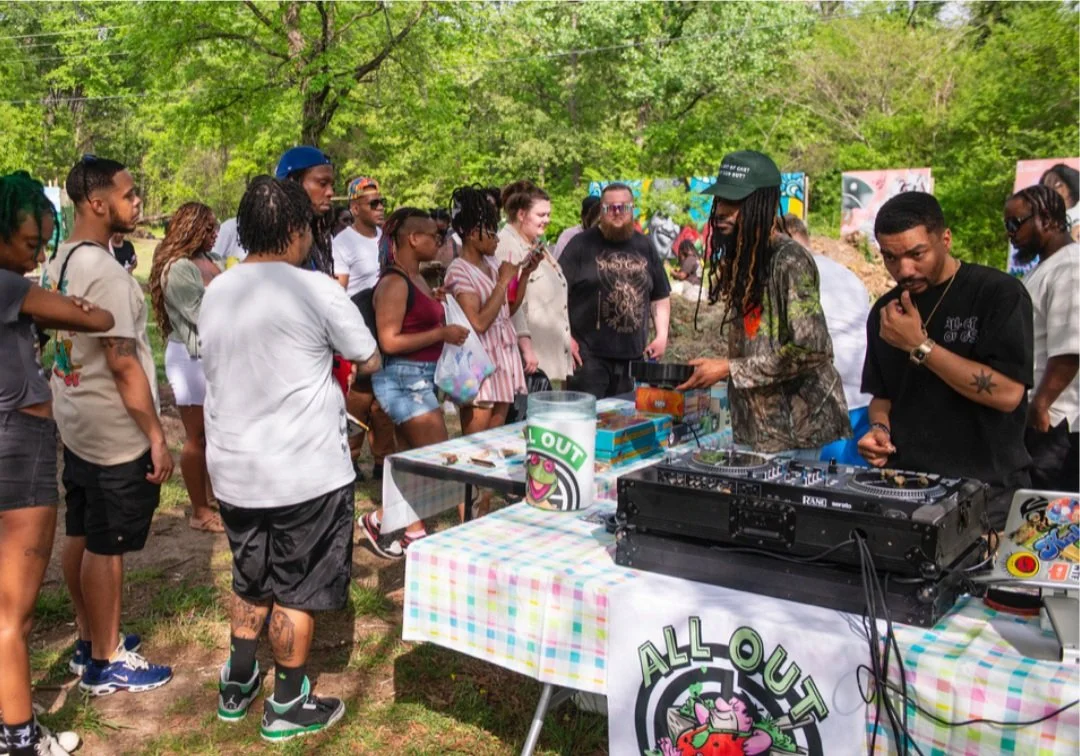 A DJ performs at an outdoor gathering with a crowd of people dancing and socializing in a lush green park.