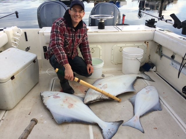 A man on a boat kneeling next to two large halibut fish with a gaff scoop, on a boat deck with fishing gear and water in the background.