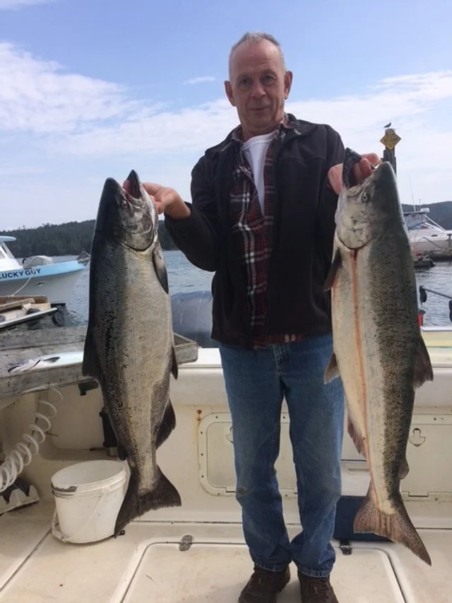 Man on a boat holding two large fish, with boats and water in the background.