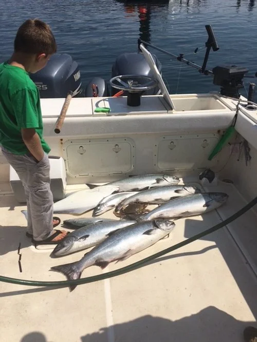 A young boy standing on a boat with caught fish laid out on the deck, near a water body with boat motors in the background.