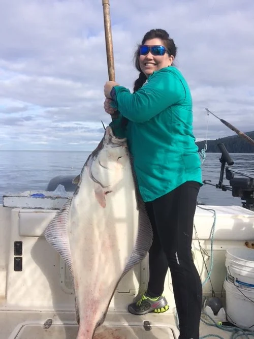 Woman in a teal jacket and black pants holding a large halibut on a boat, with water and cloudy sky in the background.