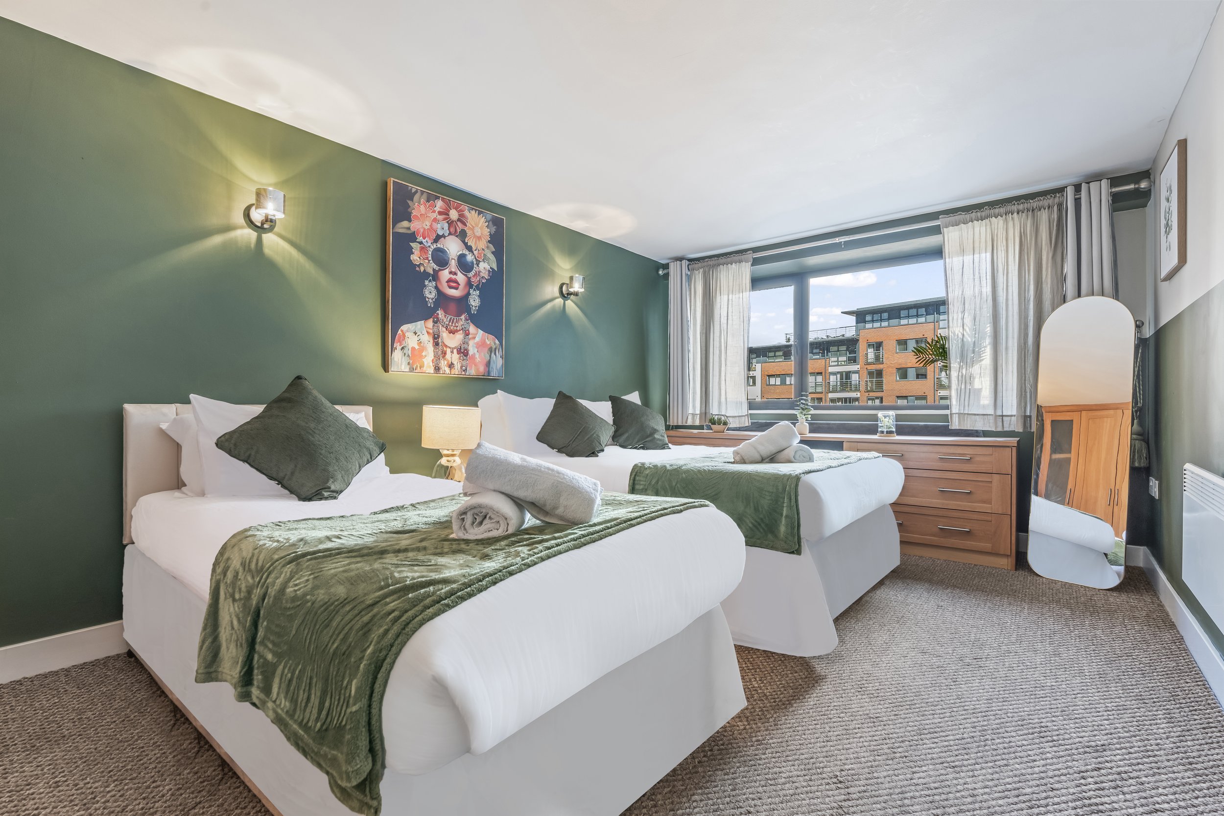 A hotel room with two beds, each with white linens and green velvet blankets, placed in front of a dark green accent wall. There is a framed artwork of a woman with flowers in her hair hanging above the beds. Light fixtures are mounted on the wall, and a large window with cream curtains allows natural light into the room.