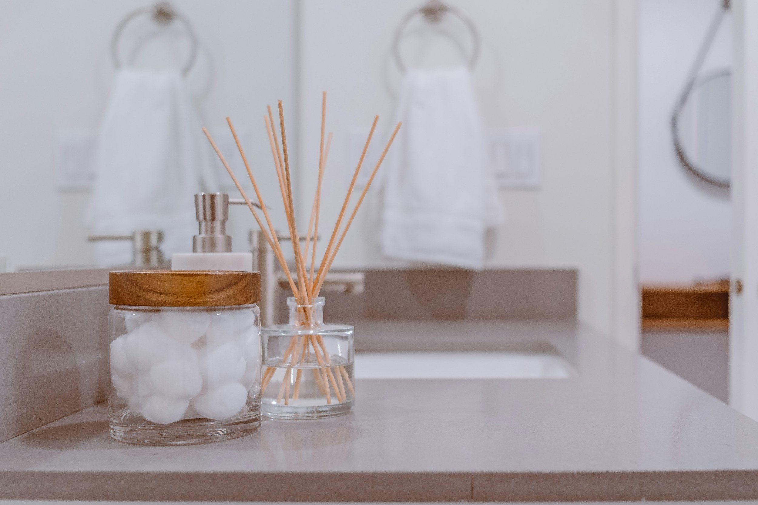 Bathroom countertop with cotton balls in a glass jar with a wooden lid, a reed diffuser, and soap dispenser, with white towels hanging on the wall in the background.