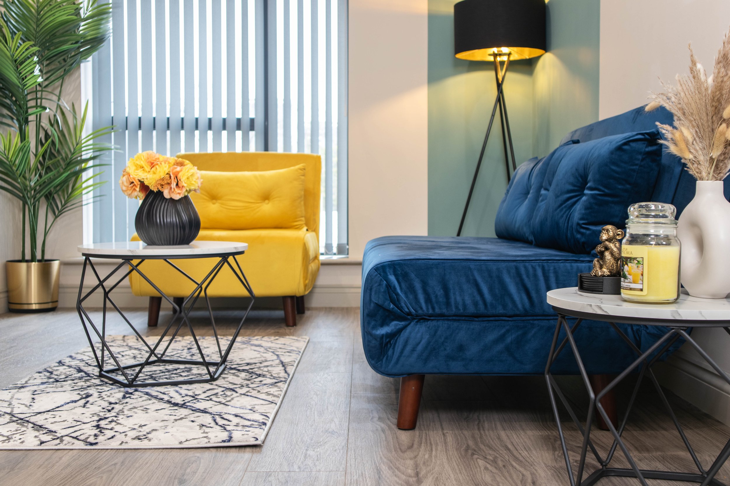 Living room with a yellow armchair and a blue velvet sofa, a white marble coffee table with a black vases with flowers, a potted plant, and a side table with a candle, a small gold bear figurine, and a white vase with dried Pampas grass. There is a f