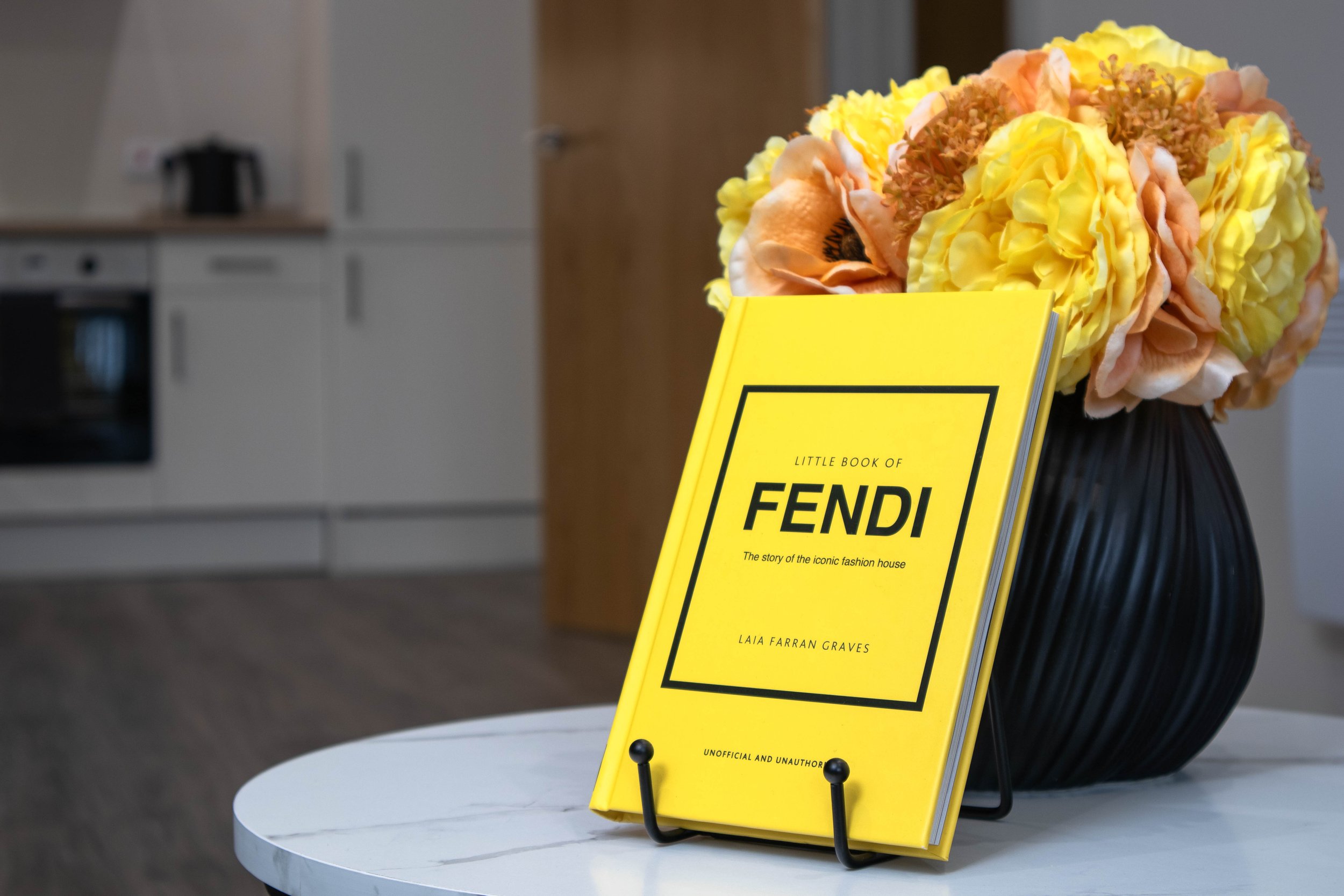A yellow book titled "Little Book of Fendi" on a stand, placed on a white marble table with a black vase holding peach and yellow flowers in the background.