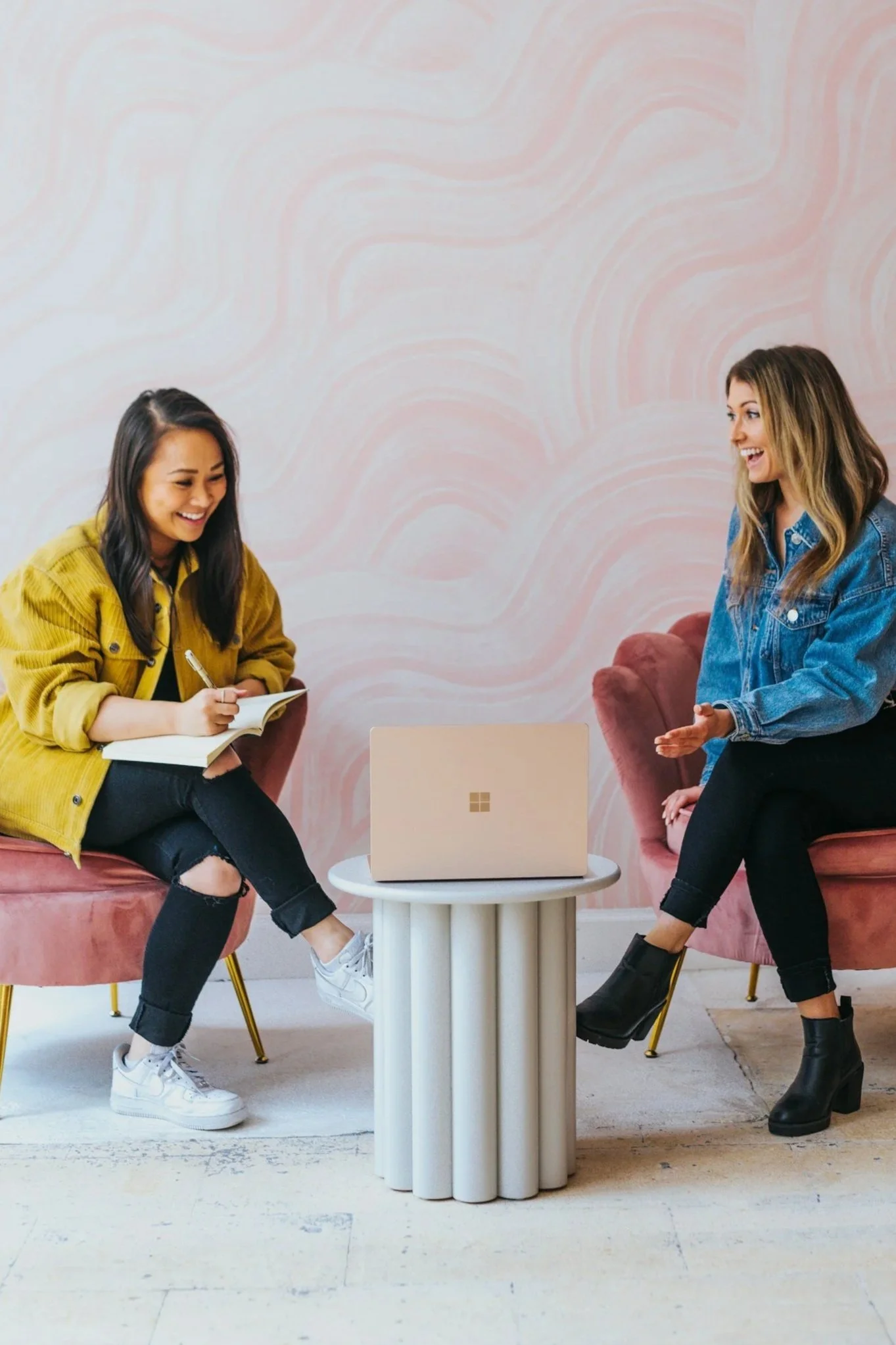 Two women sitting on pink velvet chairs talking and smiling, with a laptop on a round white table between them, in a room with pink swirled wall art.