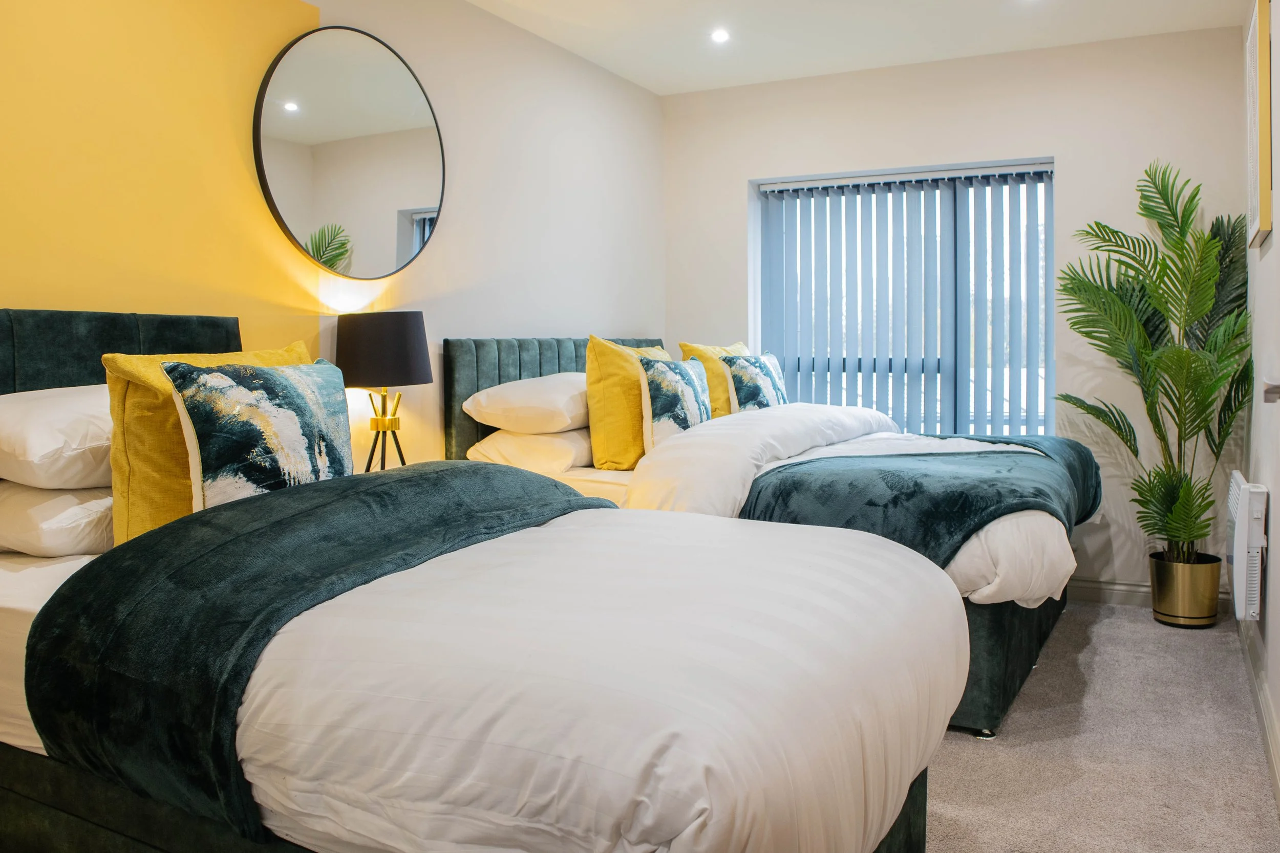 Modern bedroom with two beds, dark green velvet headboards, white bedding, yellow and patterned pillows, a large round mirror on a yellow accent wall, a black bedside lamp, a window with blue vertical blinds, a tall green potted plant, and neutral-colored walls and carpeting.