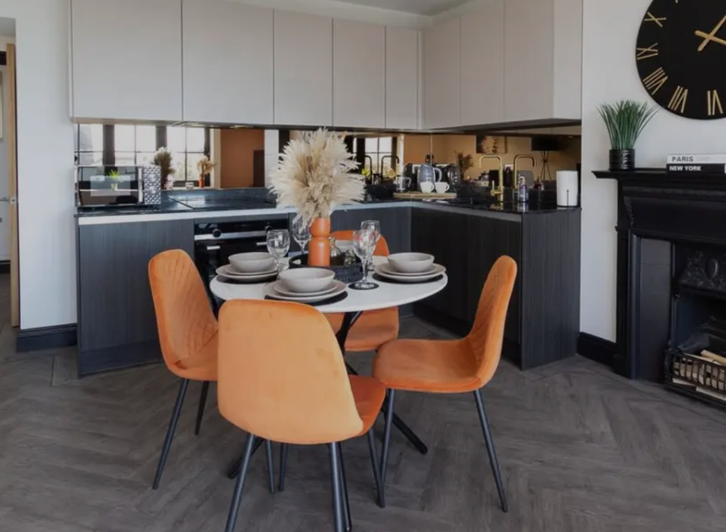 View of a modern kitchen and dining area with a round table set with plates, glasses, and a vase with dried pampas grass, surrounded by four orange chairs, a black fireplace, and a large clock on the wall.