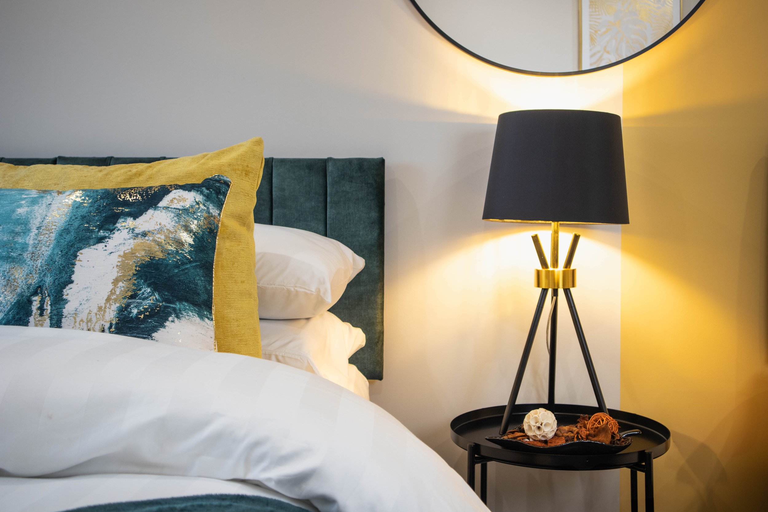 Bedroom with a bed, decorative pillows, a bedside table, a black and gold lamp, and a round mirror on the wall.