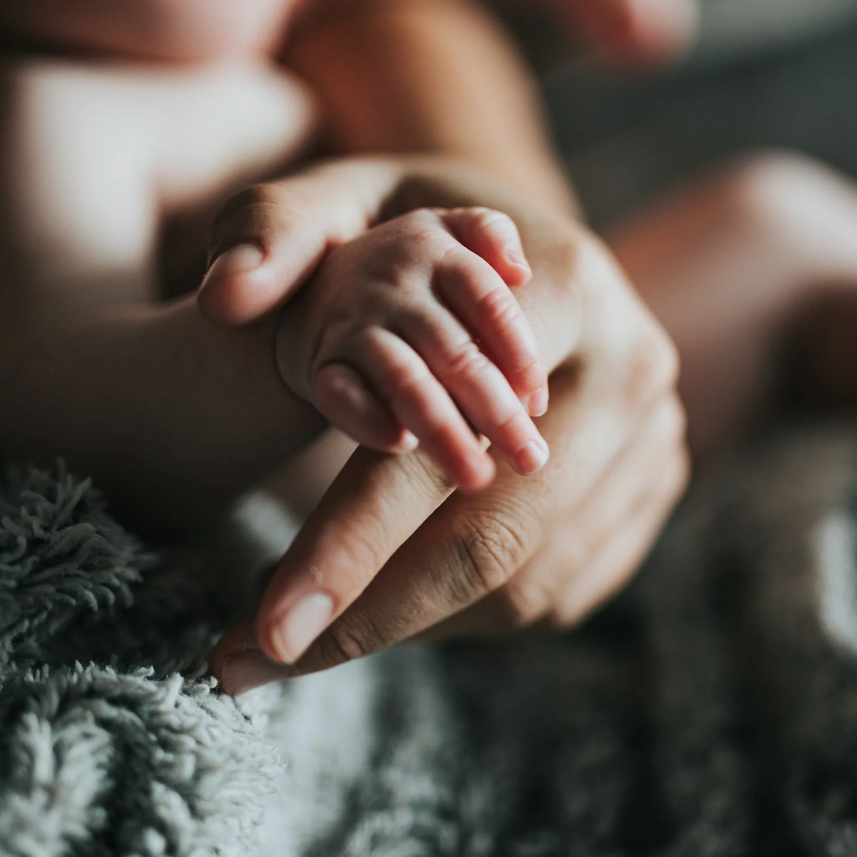 Close-up of a tiny baby's hand gripping an adult's finger, with a soft textured blanket in the foreground.