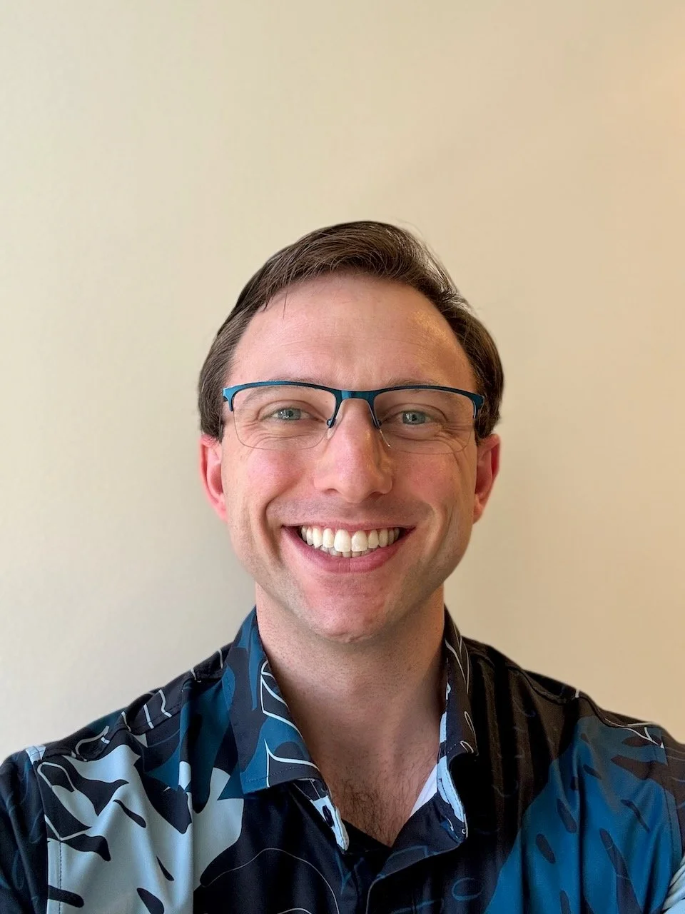 Close-up of a smiling man with glasses, wearing a black and blue patterned shirt, against a plain light background.