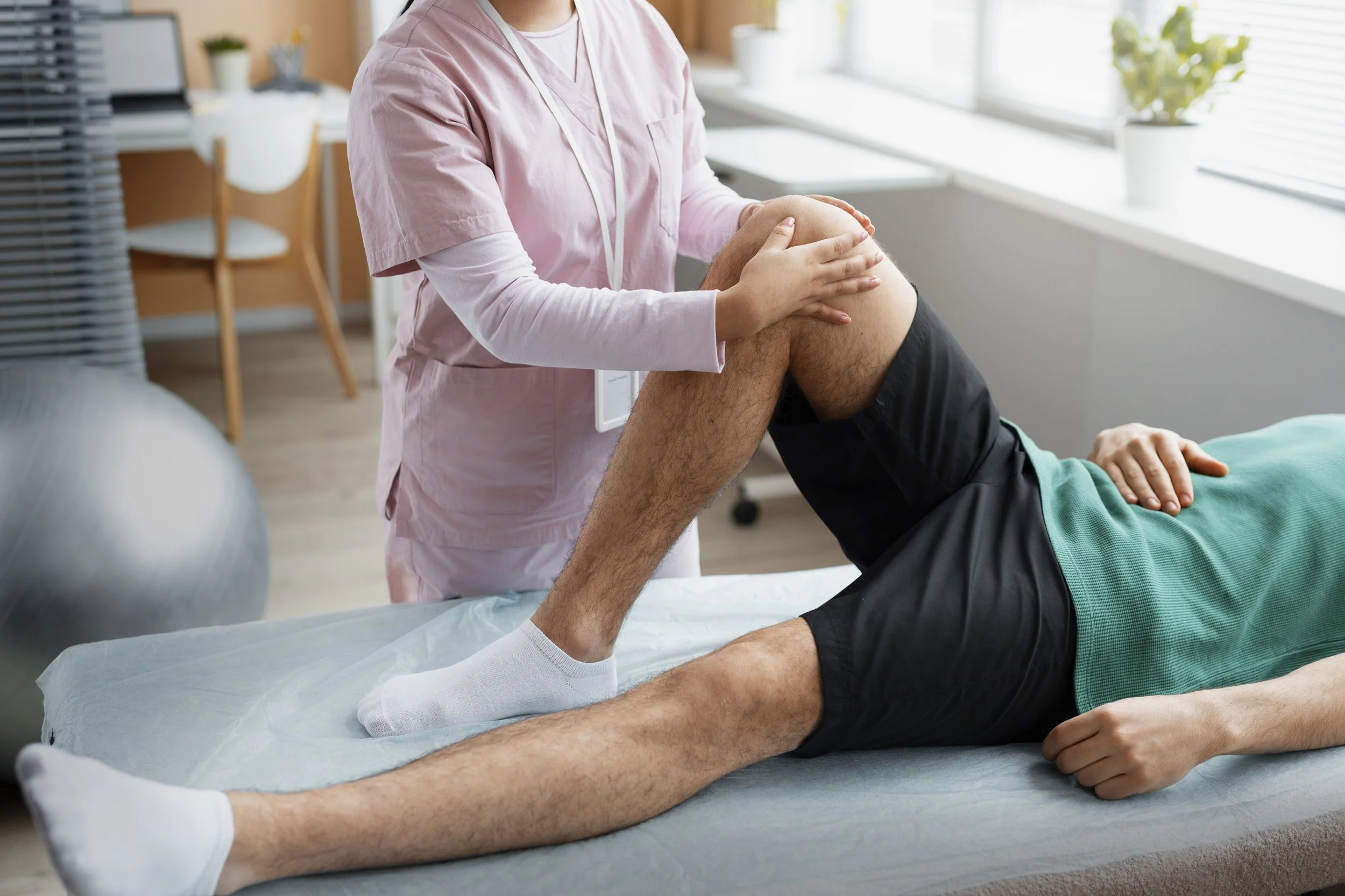 Physical therapist helping patient with leg exercises on examination table in clinic. Therapist in pink uniform supporting patient's knee, who is lying down with hand on stomach.