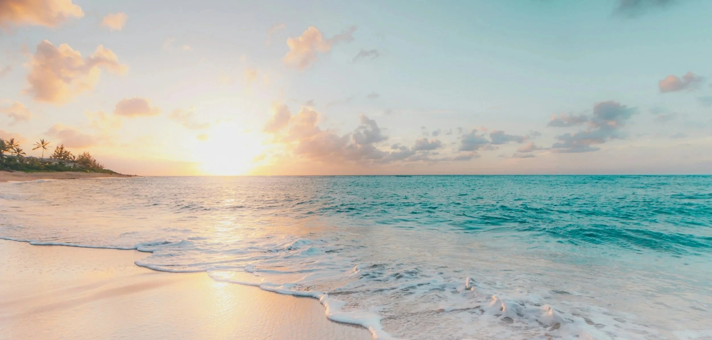 A beach scene at sunset with the sun near the horizon, calm turquoise ocean water, a sandy shoreline with small waves, and a partly cloudy sky with pink and orange hues.