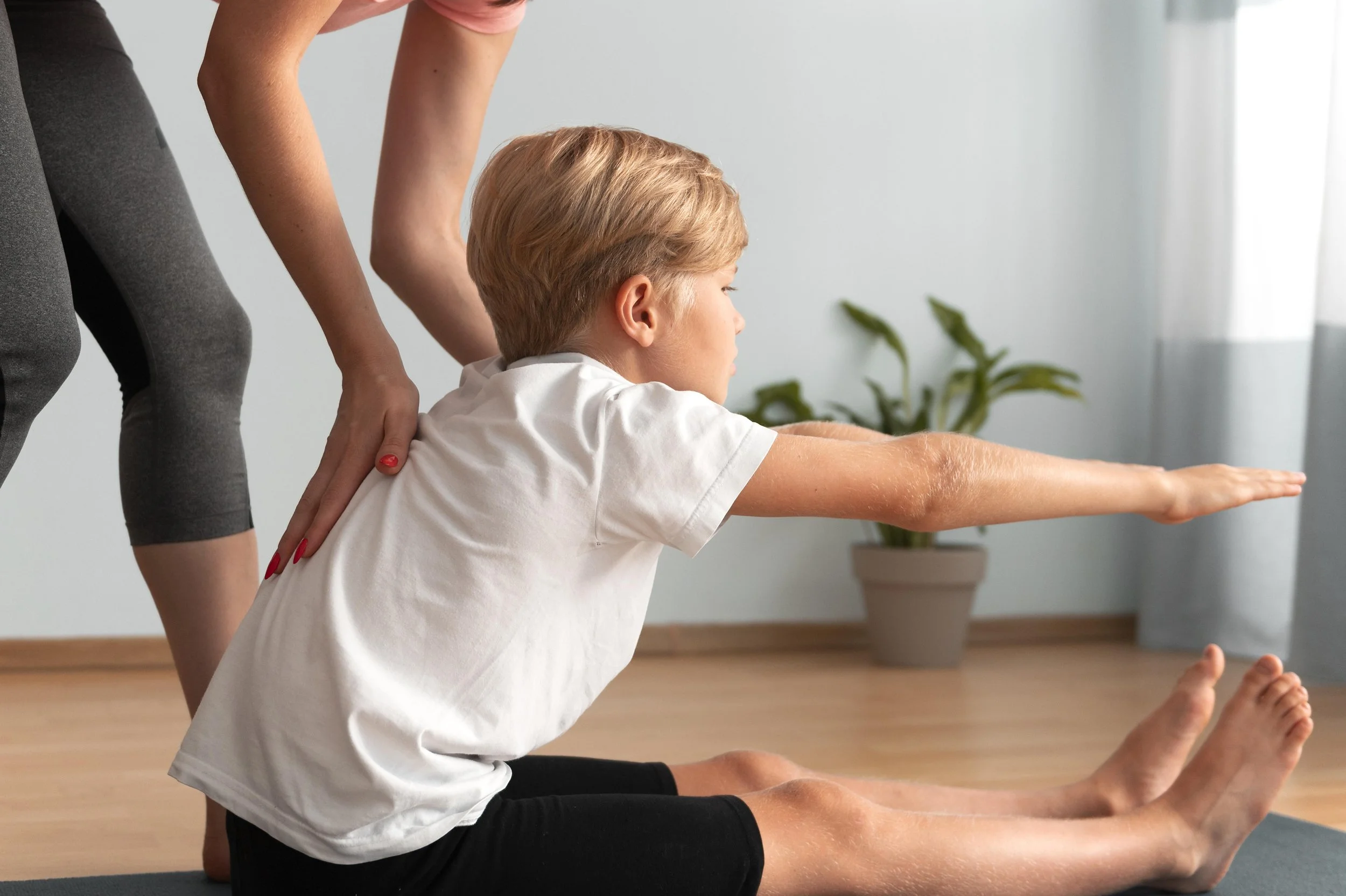 A boy with blonde hair doing yoga stretches with assistance from a woman, possibly his instructor or parent, in a room with wooden floors and a potted plant in the background.