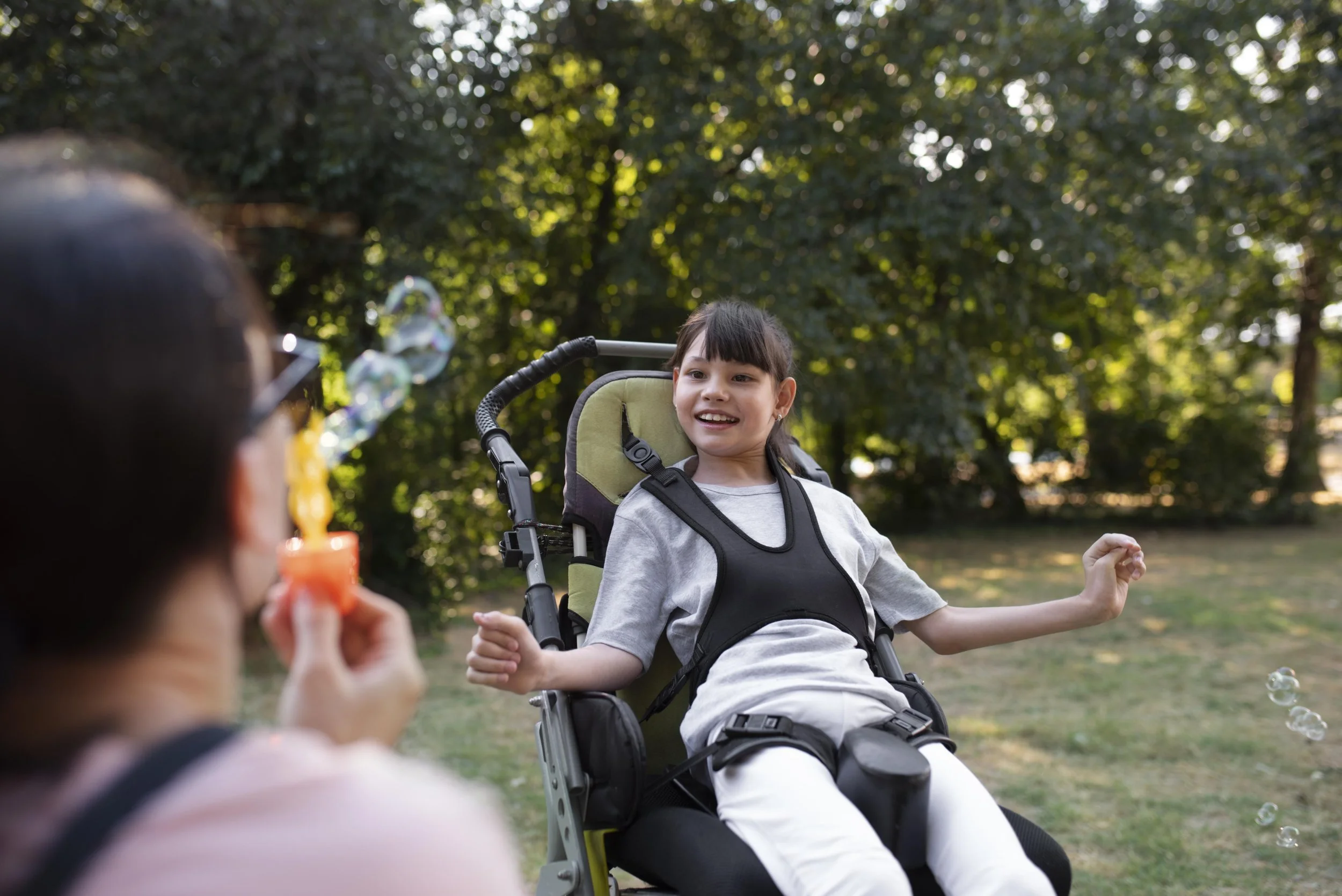 A girl in a wheelchair smiling and playing with bubbles in a park while another person, partially visible, blows bubbles toward her.