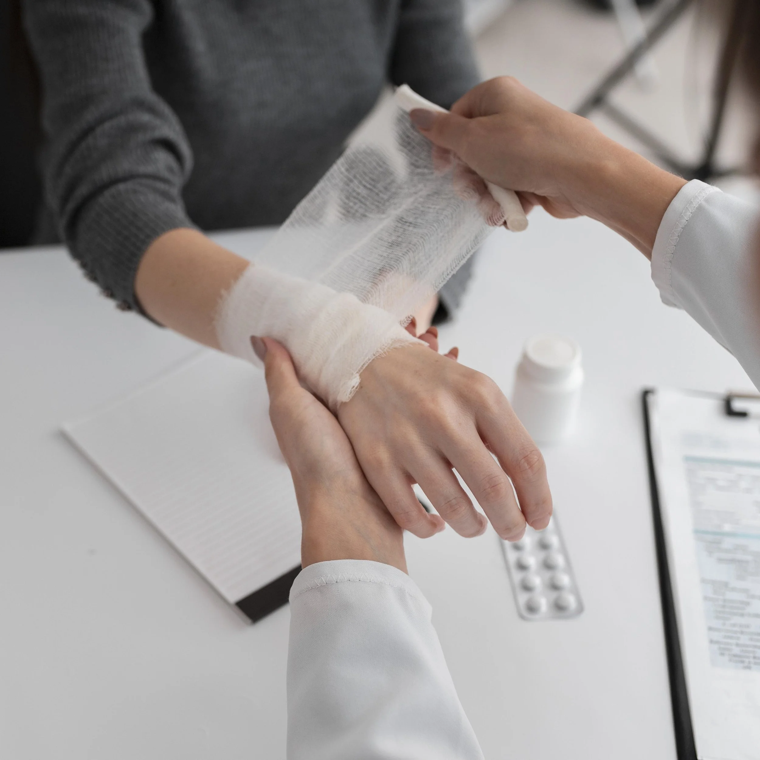 Medical professional bandages a patient's wrist in a clinical setting, with medical supplies and paperwork on the table.