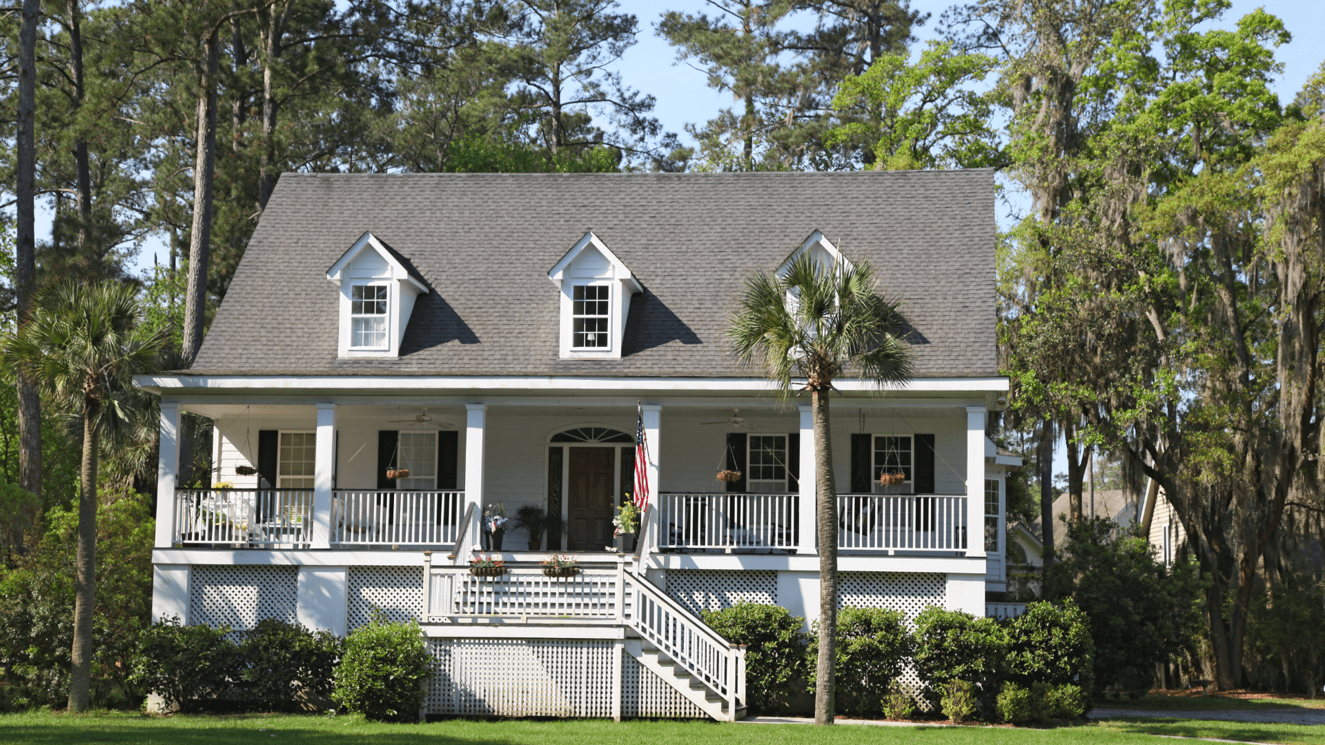 A traditional South Carolina home from the exterior. There is lush grass and Carolina Pines in the background, palm in front of elevated first floor. Full length porch.