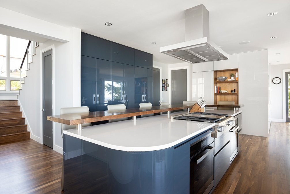 Modern kitchen with blue cabinets, wooden accents, and a white countertop island. Features built-in appliances, shelves with decor, and a staircase nearby.