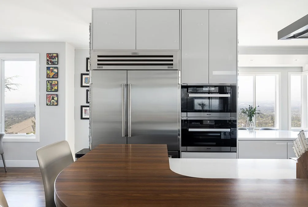 Modern kitchen with stainless steel refrigerator and built-in microwave, white cabinets, and large windows showing an outdoor view.