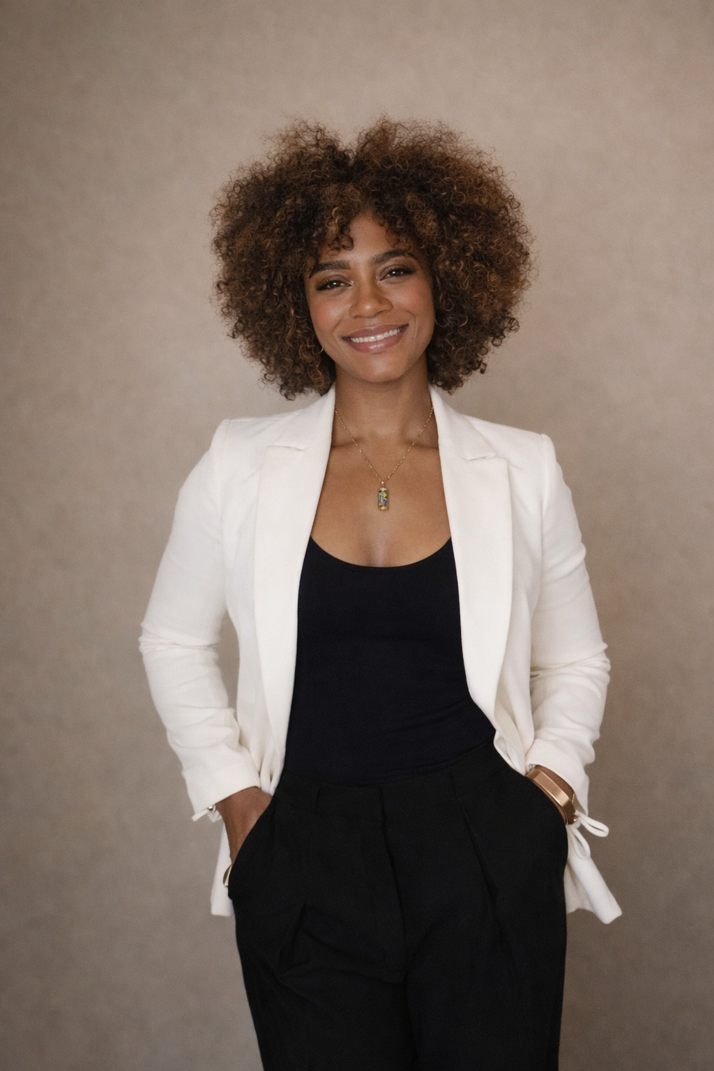 A woman with curly brown hair wearing a white blazer, black top, and black pants, smiling and posing with her left hand in her pocket against a plain background.
