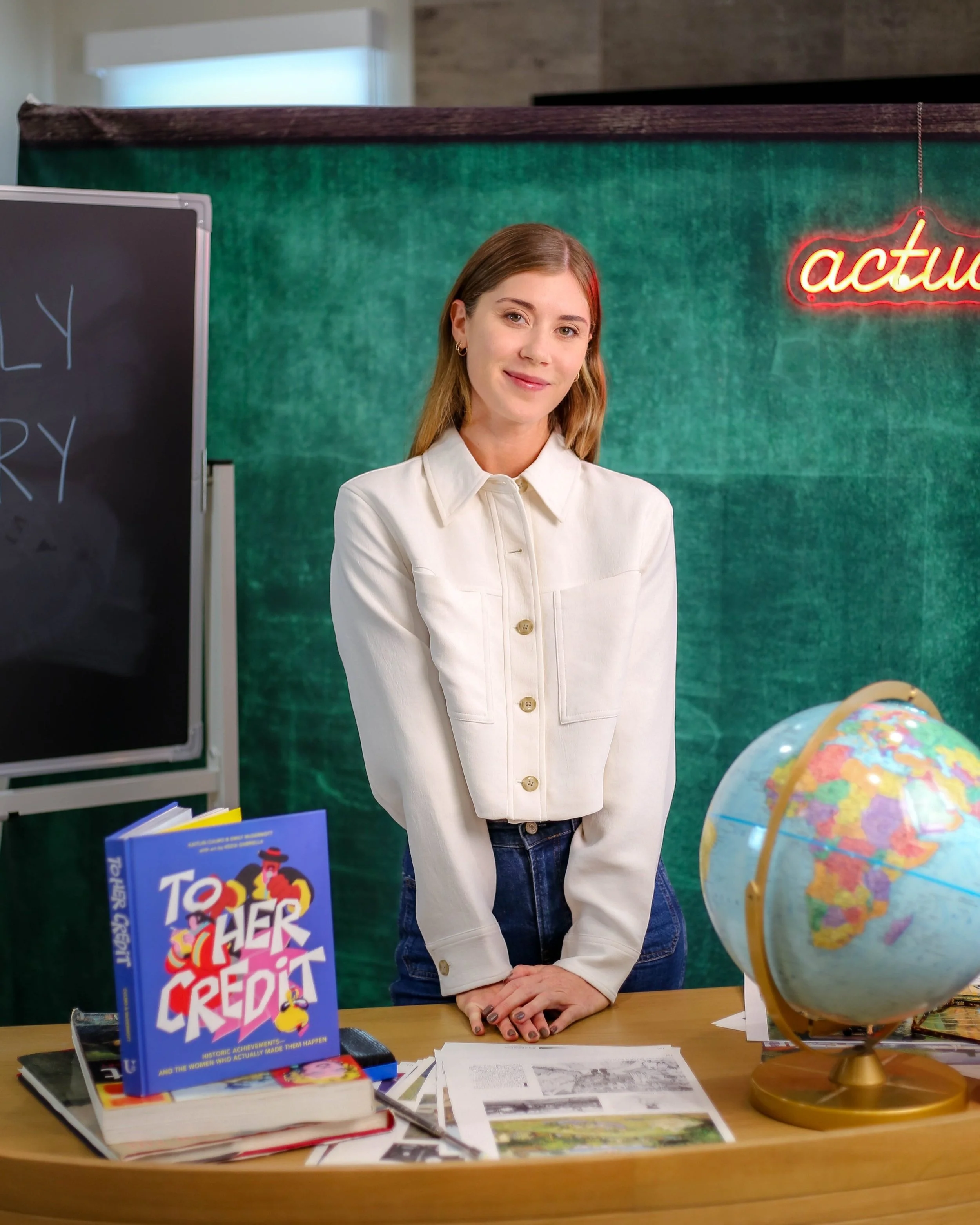 A young woman standing behind a wooden teacher's desk in a classroom, with a globe and books, in front of a green chalkboard and a neon 'active' sign.