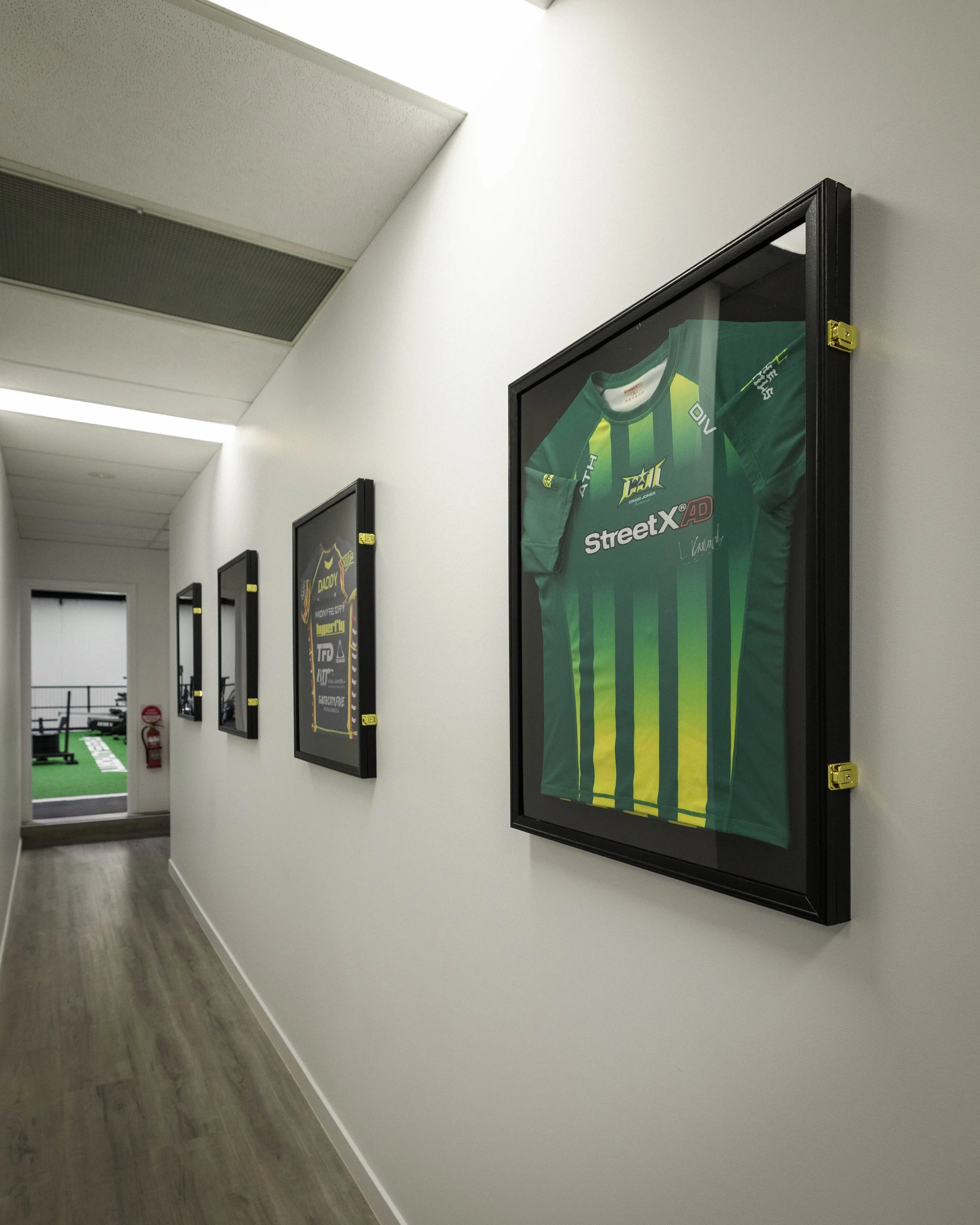 Hallway with framed sports jerseys on the wall, including a green and yellow striped football jersey, with ceiling vents and a fire extinguisher visible in the background.