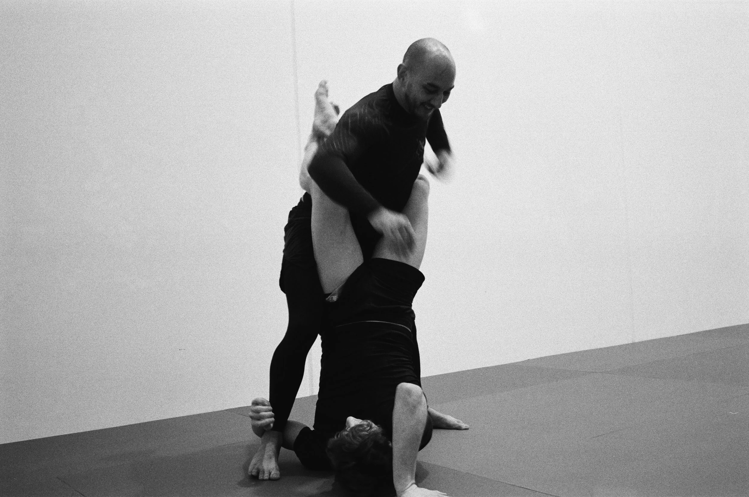 Two individuals practicing acro yoga in a gym, with one person balancing on hands and knees while the other supports and balances on their back.