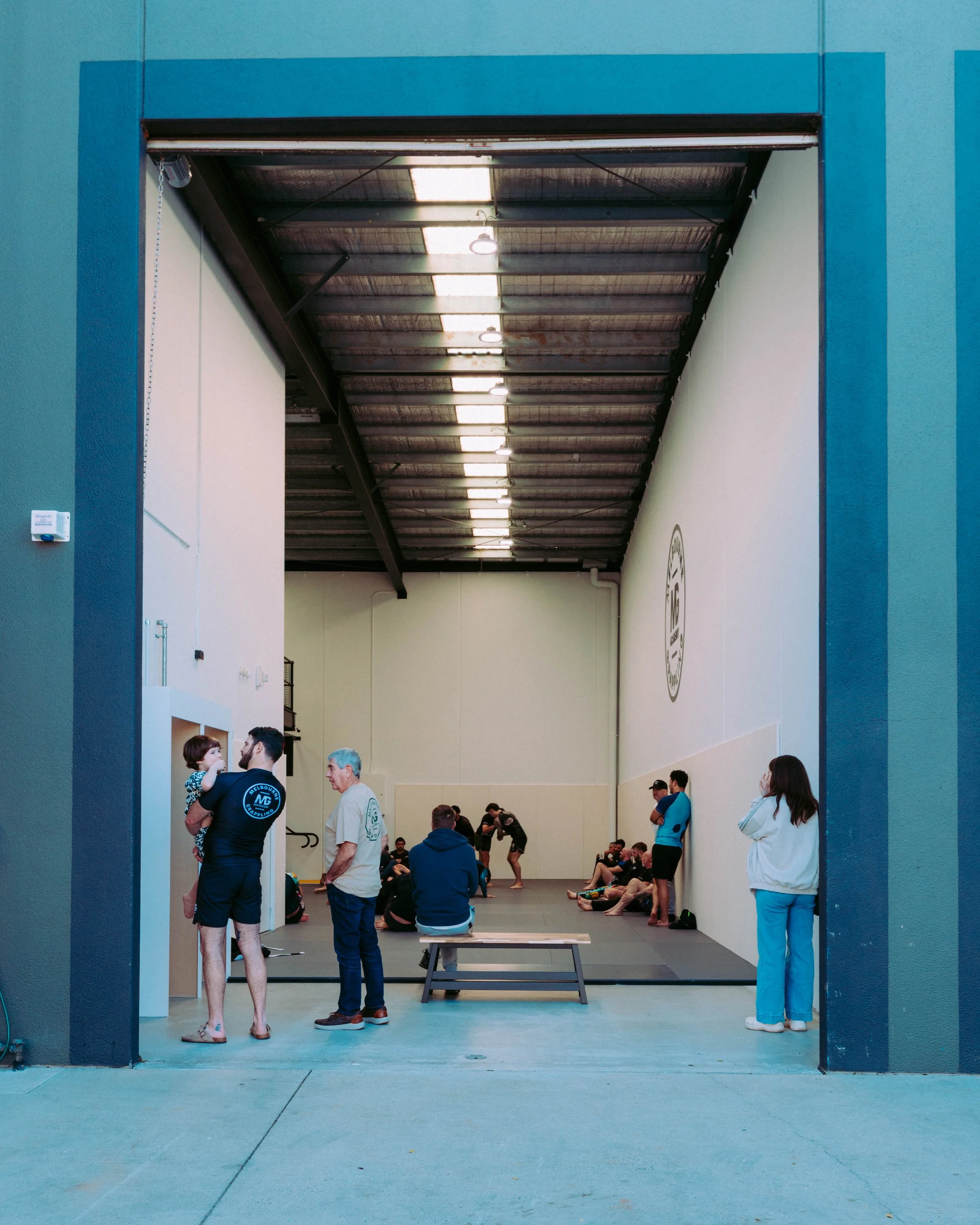 People gathering inside a large, open indoor sports facility, with some chatting, walking, or sitting on the floor. The space has high ceilings with lighting and a wall with a logo or emblem.