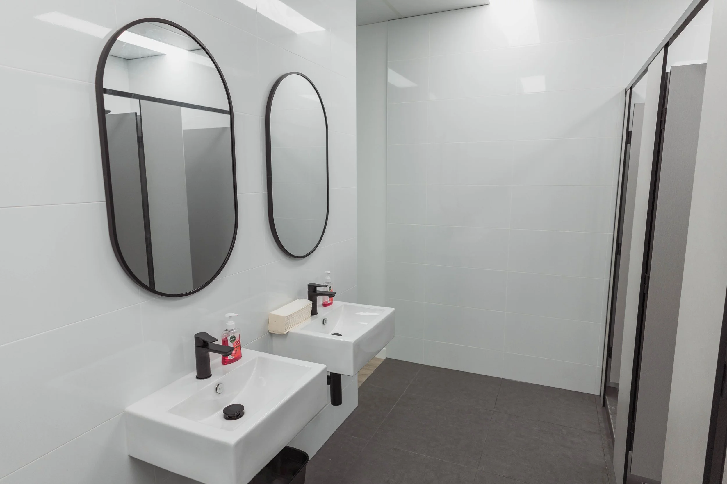 A modern public restroom with two white sinks and black faucets, oval mirrors, hand soap, and a soap dispenser on the countertop. The walls are covered with white tiles, and there are gray tiles on the floor. A partial view of a stall partition can be seen on the right.