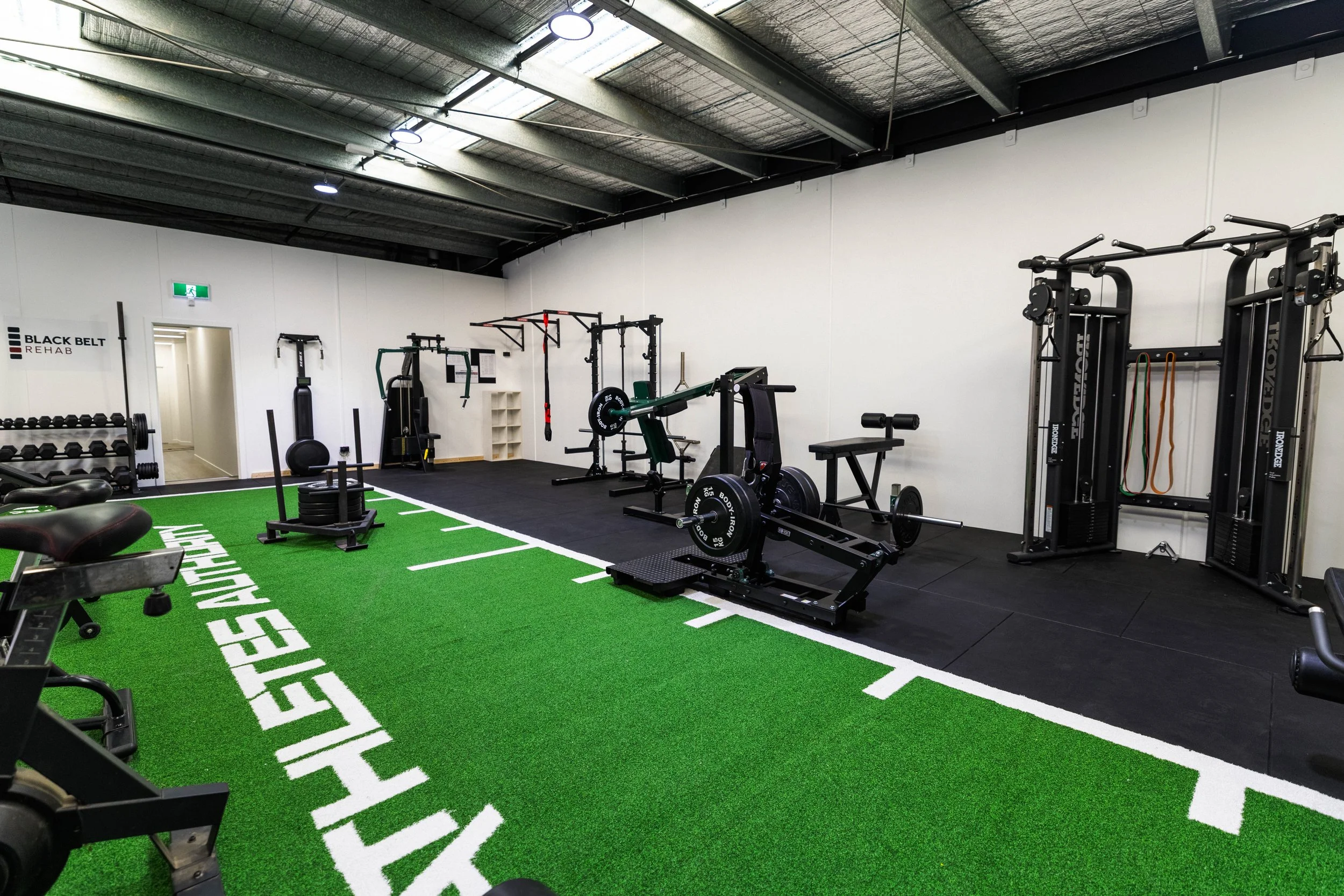 Interior of a gym with various workout equipment, including weight machines, dumbbells, and benches, and a green turf area with white markings for fitness exercises.