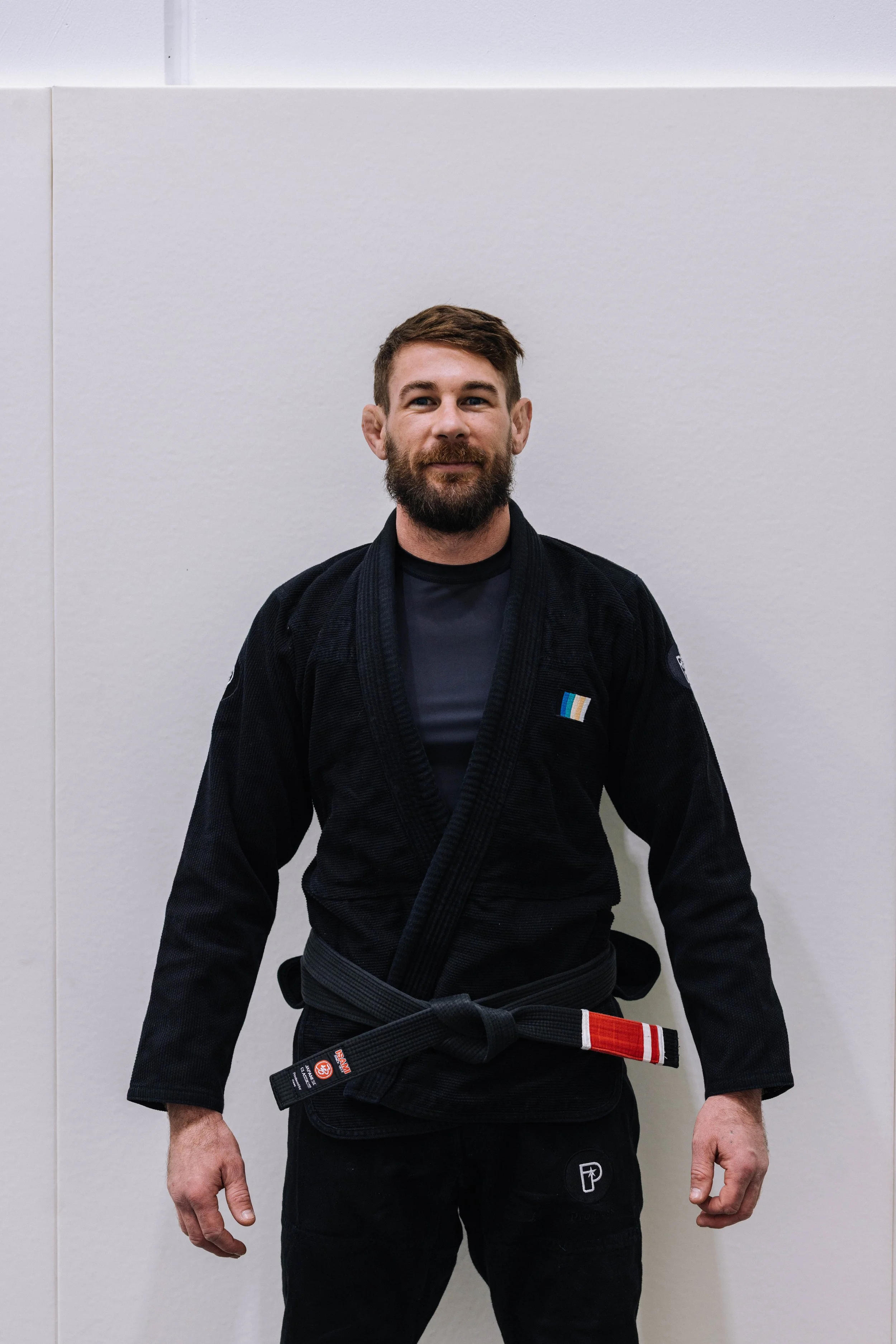 A man standing in a martial arts gi with a black belt, facing forward, against a white wall.