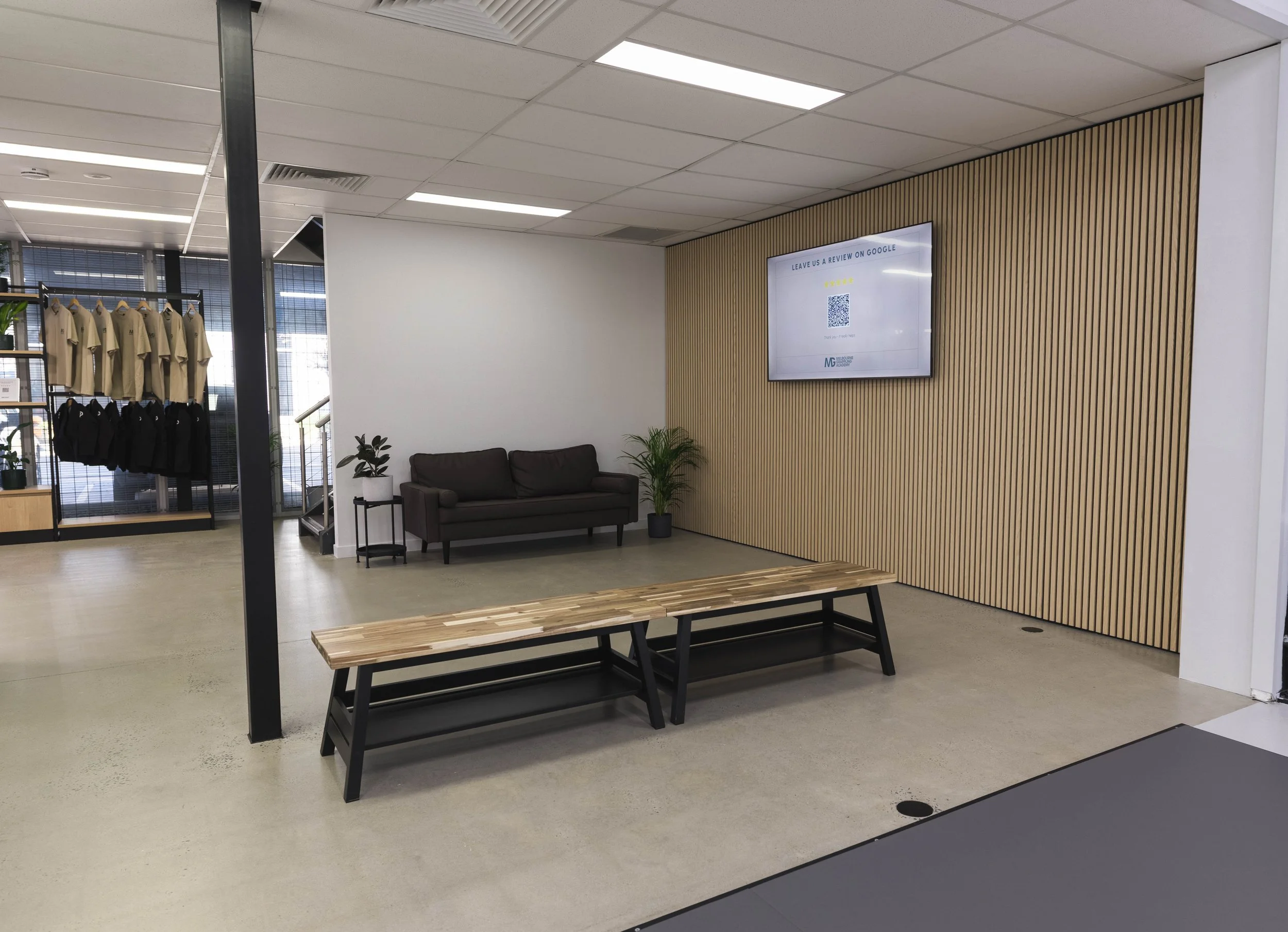 Modern reception or waiting area with a brown sofa, two potted plants, a wooden bench, a wall-mounted screen displaying a review prompt, and a clothing display with beige and black shirts.