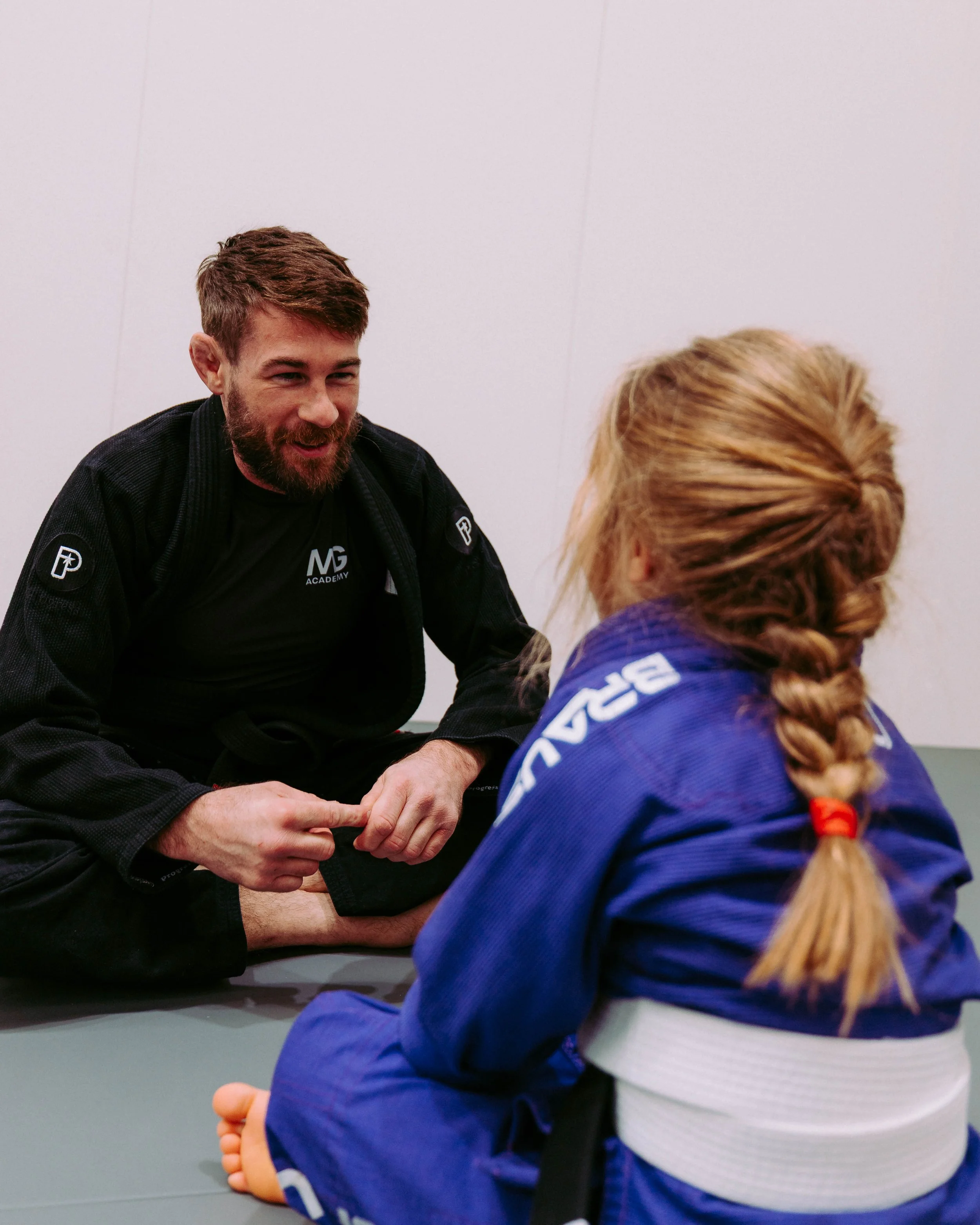 A man with a beard and short brown hair, wearing a black martial arts uniform with patches, is smiling and holding a small child's foot. The child, dressed in a blue martial arts gi with a white belt, has long blonde hair tied back with a red hair tie and is sitting at a table with the back of their head visible. They are in a room with plain white walls.