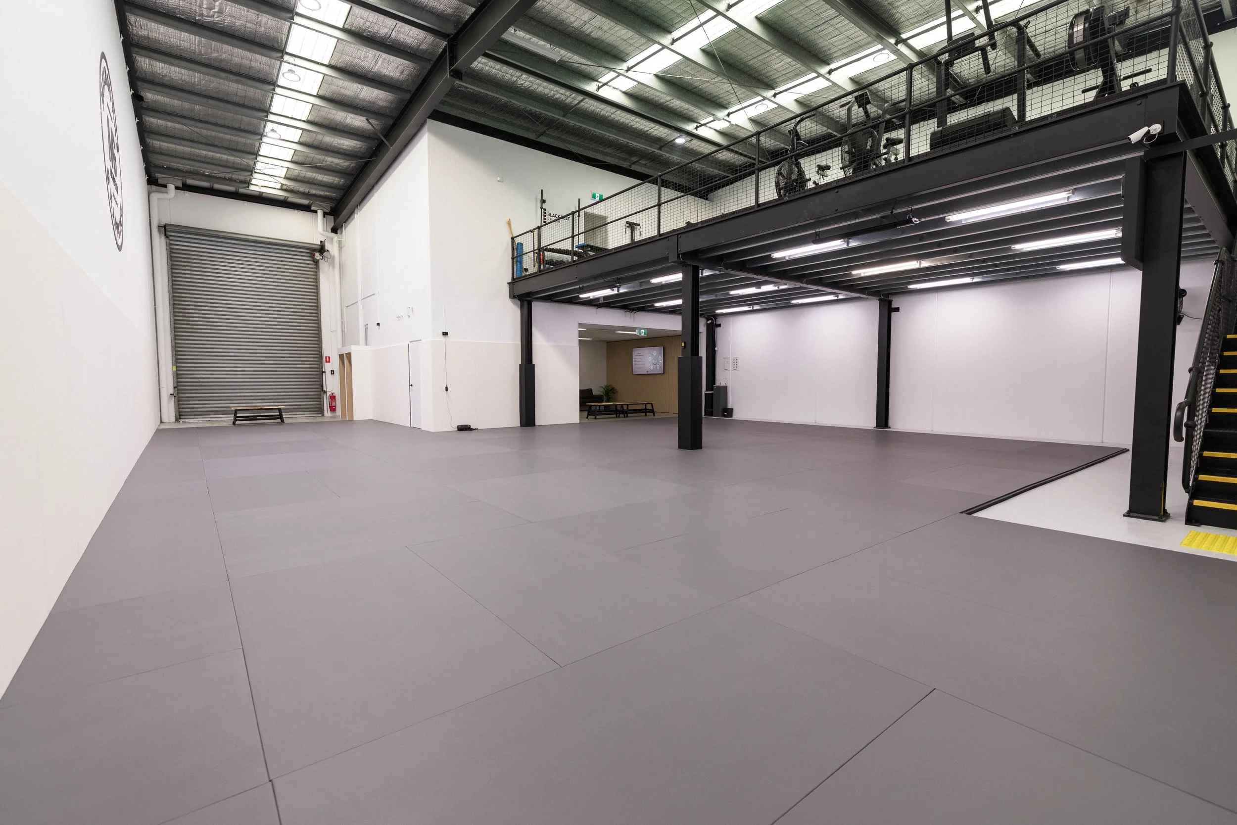 A spacious, empty indoor gym or fitness studio with gray padded flooring, white walls, a rolling metal door, and a black upper walkway with exercise bikes and equipment. The ceiling has exposed metal beams and insulation, with bright fluorescent lighting.