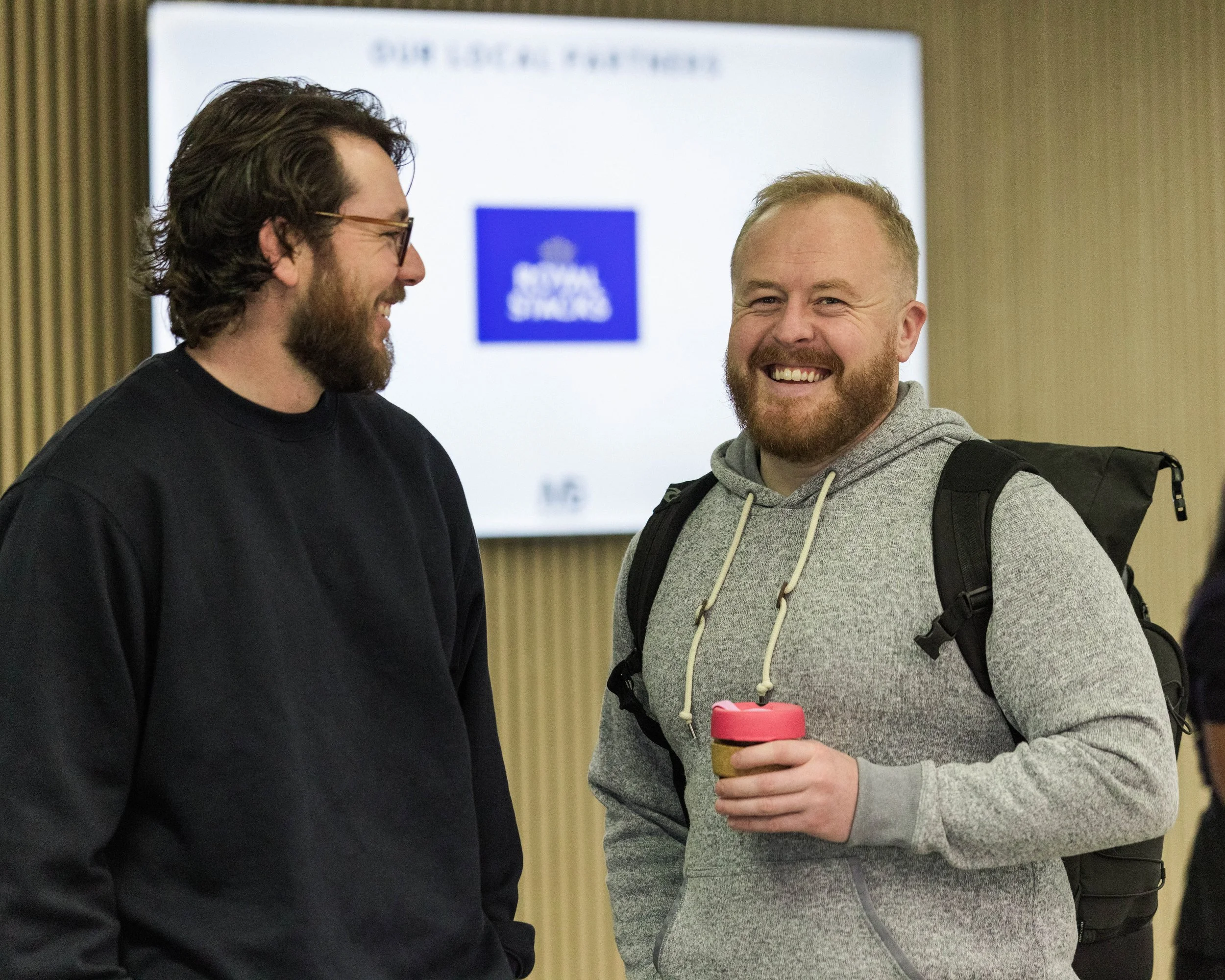 Two men smiling and talking indoors. One is holding a pink-lidded coffee cup. The man on the left has glasses and a beard, and the man on the right has a beard and a backpack. In the background, a digital screen displays a blue logo and some text.