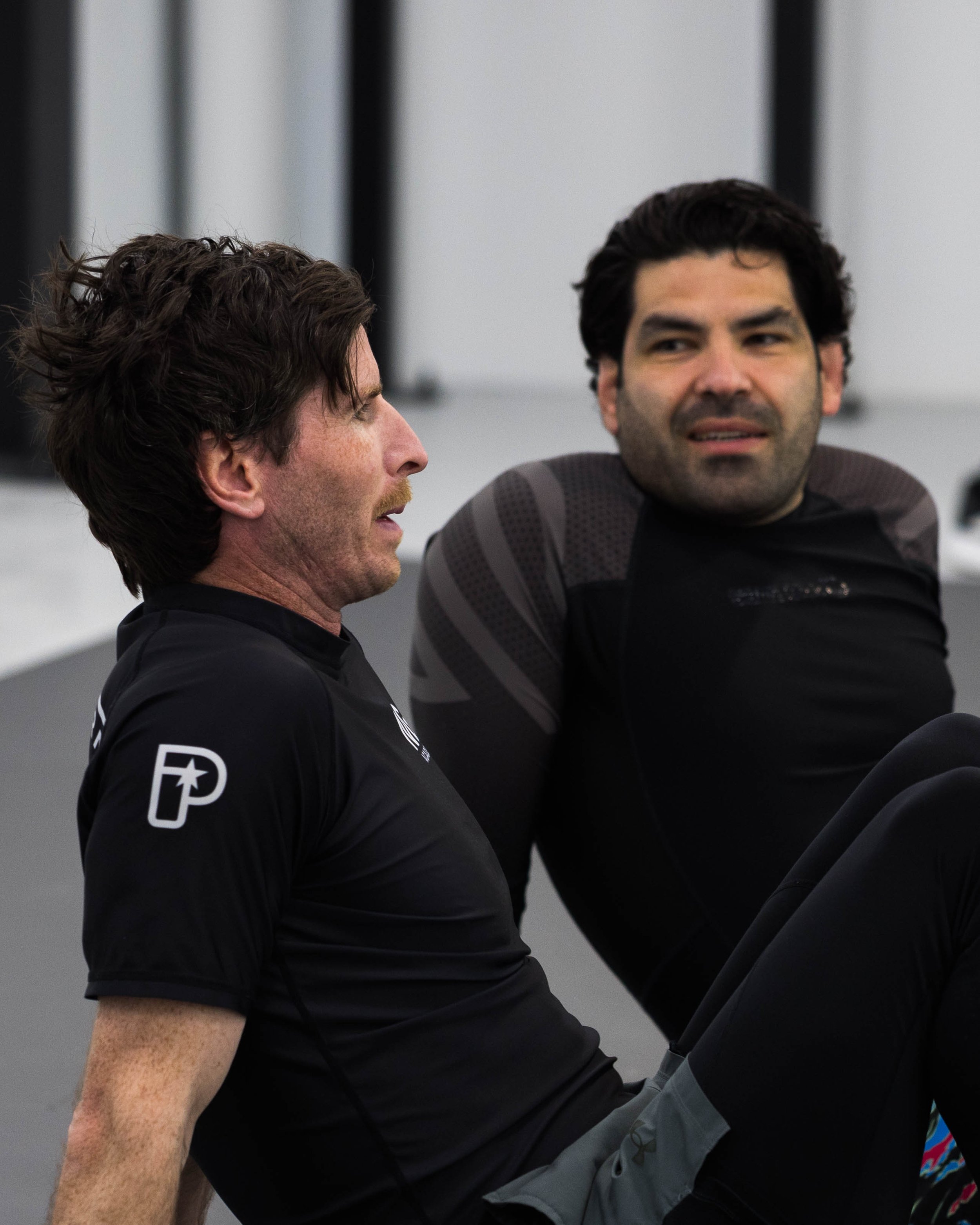 Two men sitting and talking in an indoor setting, one with brown hair and the other with dark hair, wearing sportswear.