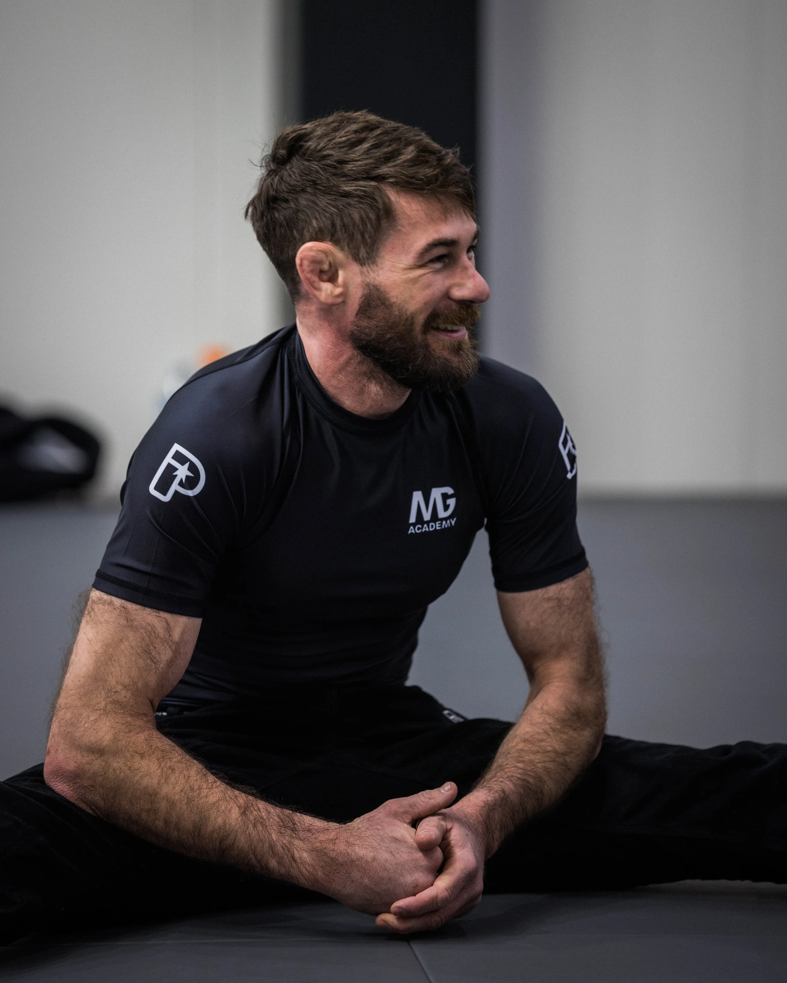 A man with a beard and short brown hair, wearing a black shirt with a white logo and the letters 'MG' on it, sitting on a black mat in a dark room. He is smiling and has his arms resting on his bent knees, with hands clasped together.