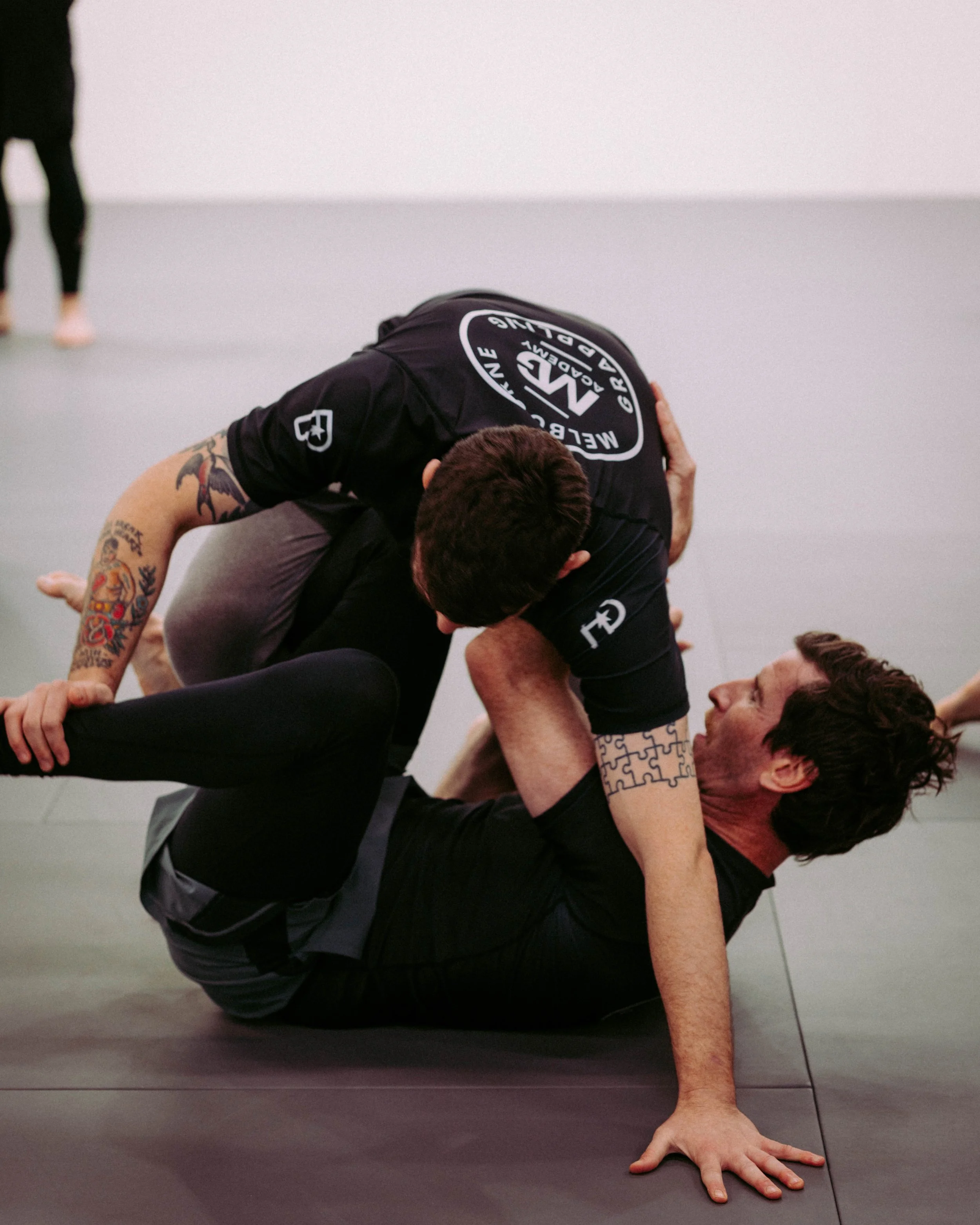 Two men practicing Brazilian Jiu-Jitsu on a gray mat, one man on top in a dominant position, and the other on the bottom gripping the top man's arm.