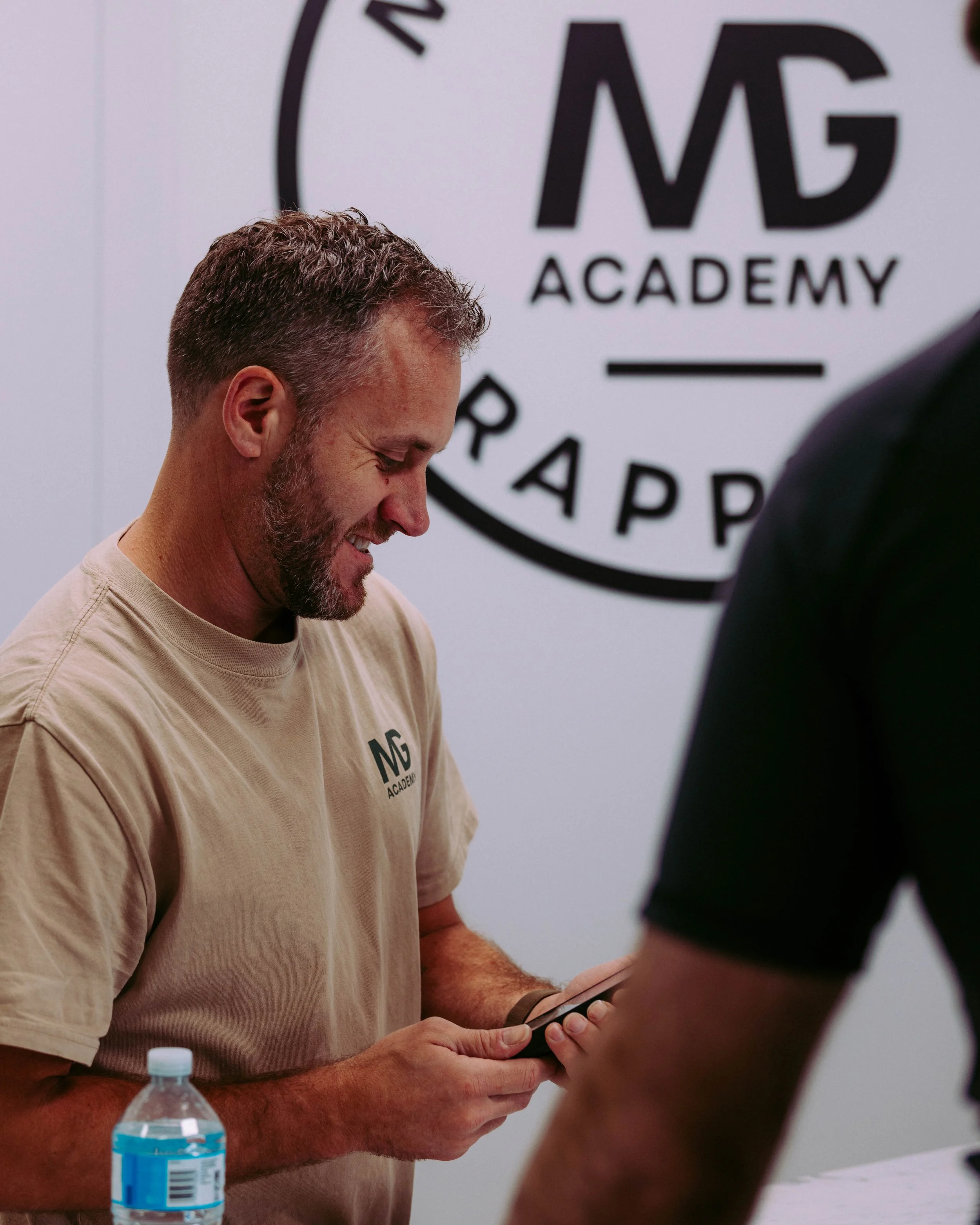 A man with short hair and a beard wearing a beige t-shirt with 'MG ACADEMY' written on it, smiling while looking at a smartphone held in his hands. A bottle of water is on the table in front of him. In the background, a wall displays a partial logo with the text 'MG ACADEMY'.