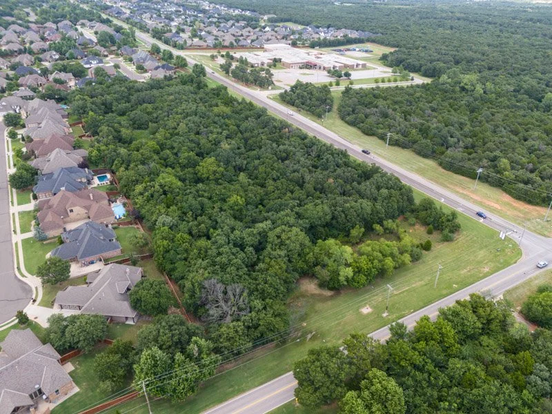 Aerial view of a large wooded area with a property boundary marked in yellow, adjacent to a residential neighborhood and a major road with traffic.