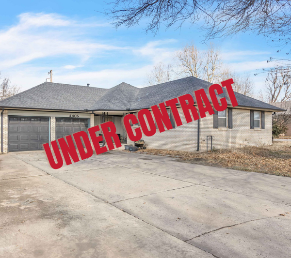 A single-story house with a gray shingle roof, beige brick exterior, black window shutters, and a double garage with black doors. The driveway is concrete with some cracks, and there are a few potted plants and garden debris near the house. Bare trees and neighboring houses are visible.