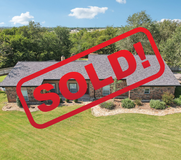 Single-story house with brick and beige siding, front yard with green grass and landscaped garden beds, blue sky with clouds, and trees in the background.