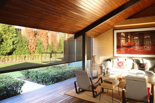 Living room with large window overlooking outdoor garden, featuring a beige sofa with patterned cushions, a wooden armchair, coffee table, and wall art.