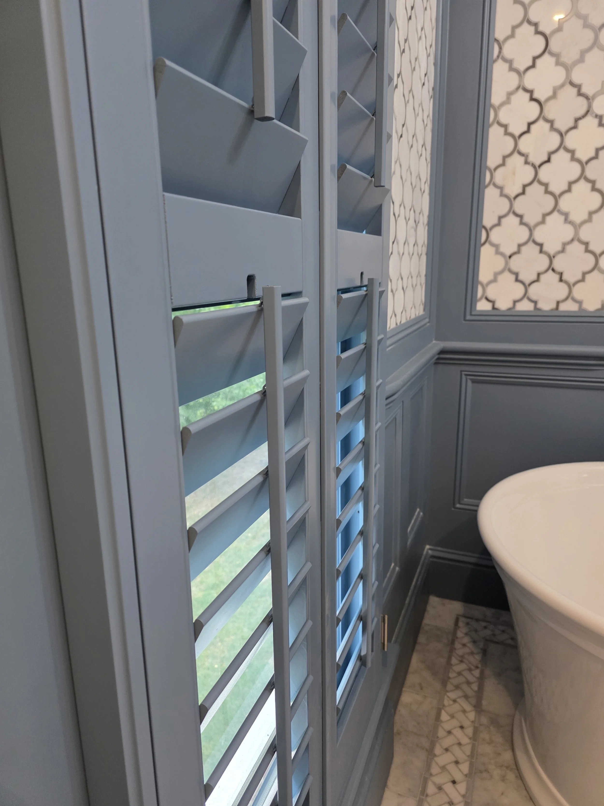 A bathroom with gray wooden window shutters and patterned wallpaper next to a white toilet.