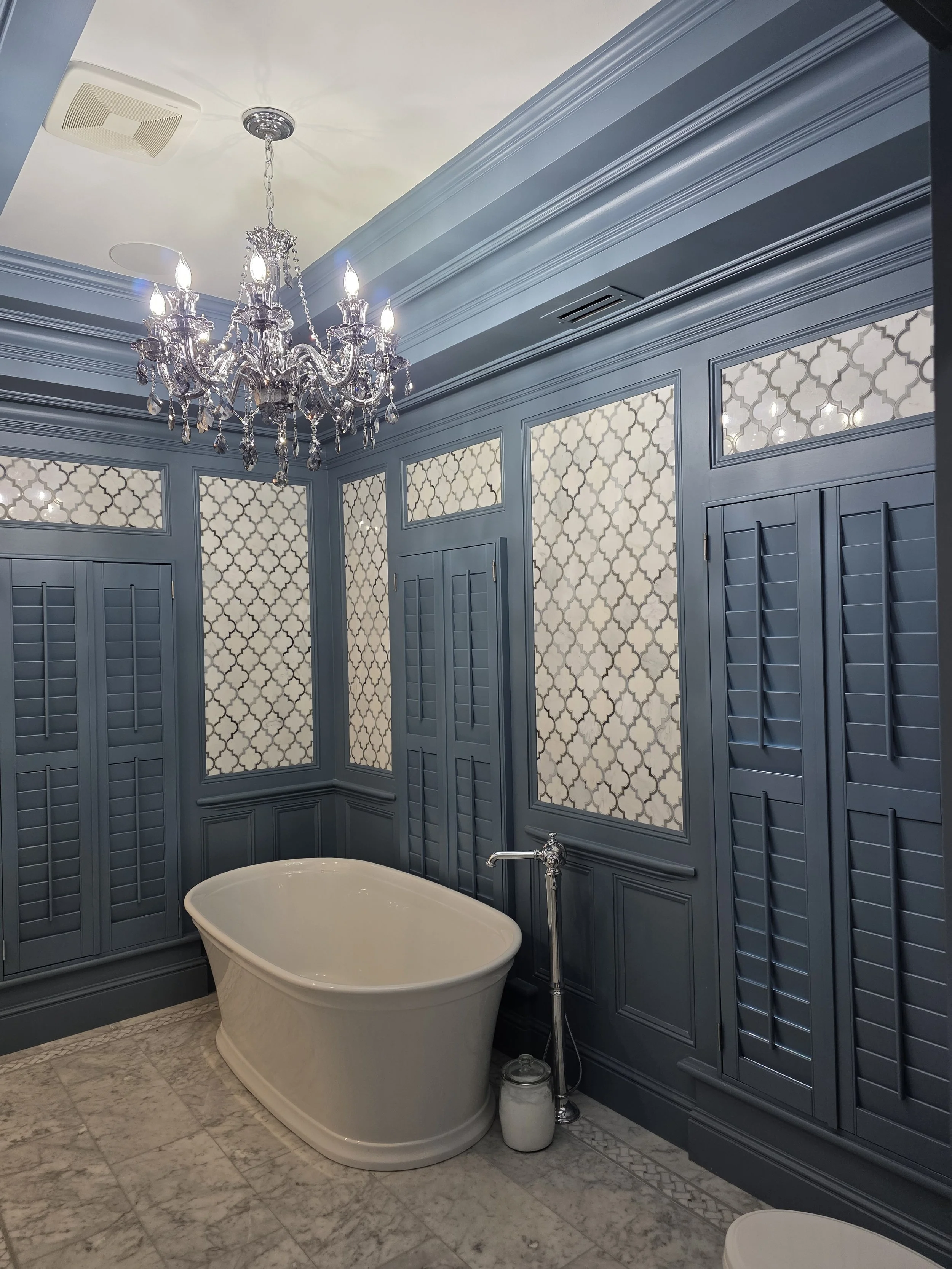 Elegant bathroom with a standalone bathtub, blue wainscoting and patterned wallpaper, crystal chandelier, marble floor, and built-in shutters.