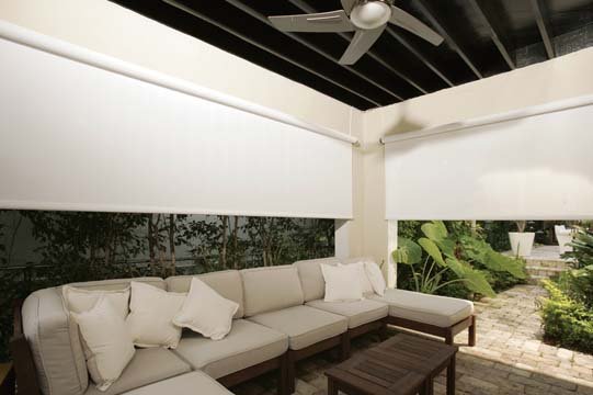 Outdoor patio area with beige sectional sofa, white pillows, wooden coffee table, ceiling fan, and large green plants outside, partially enclosed by white walls and window shades.
