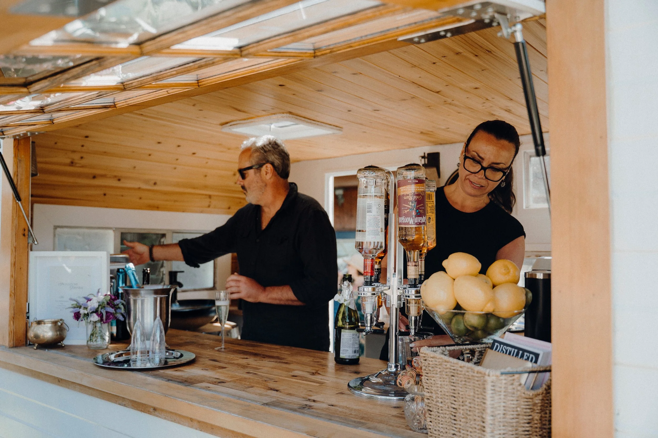 A man and woman working behind a wooden bar, with the man reaching for something and the woman looking down, surrounded by bar supplies and lemons.