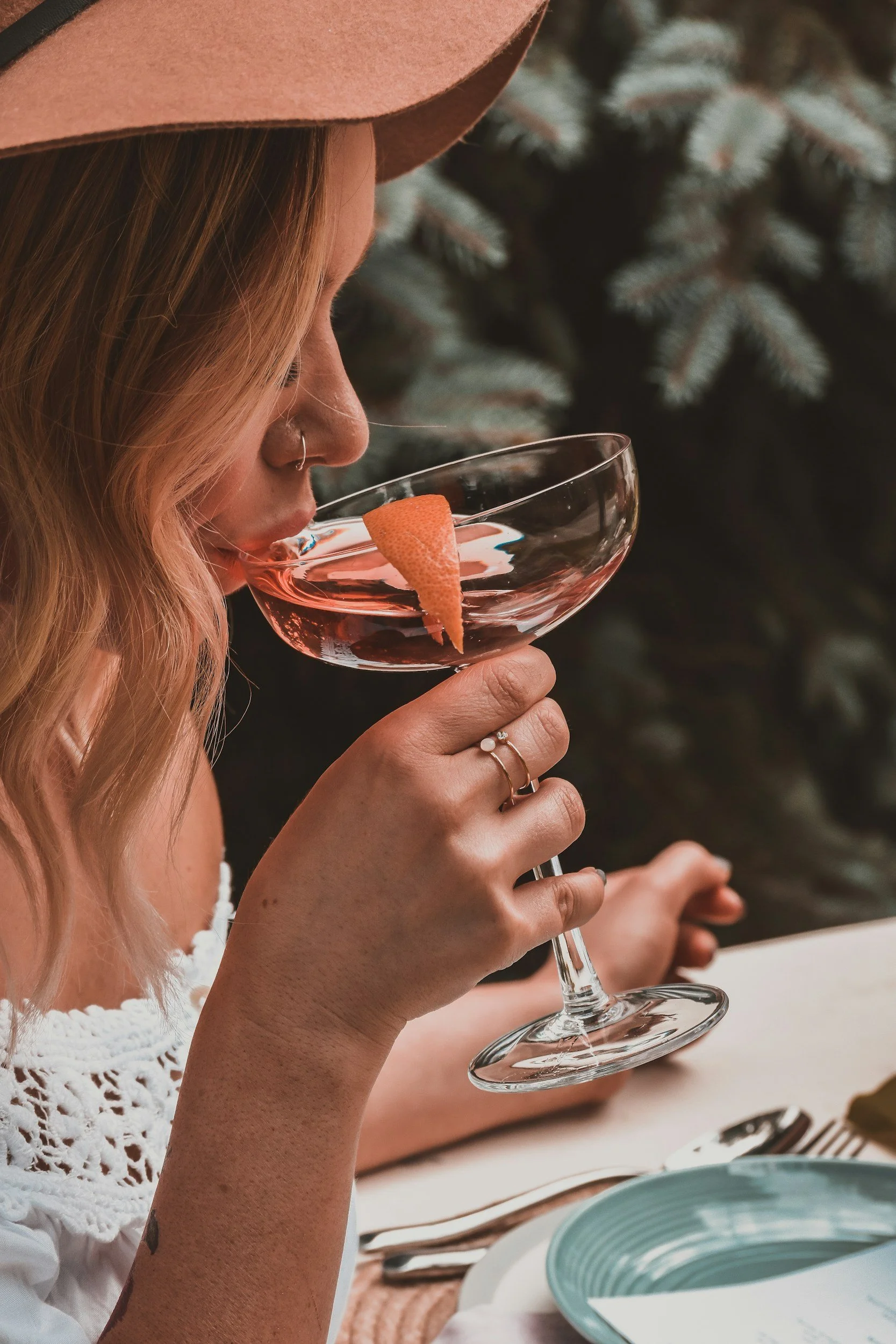 A woman with light-colored hair and a hat is drinking a pink cocktail garnished with an orange peel, seated at a table outdoors with greenery in the background.