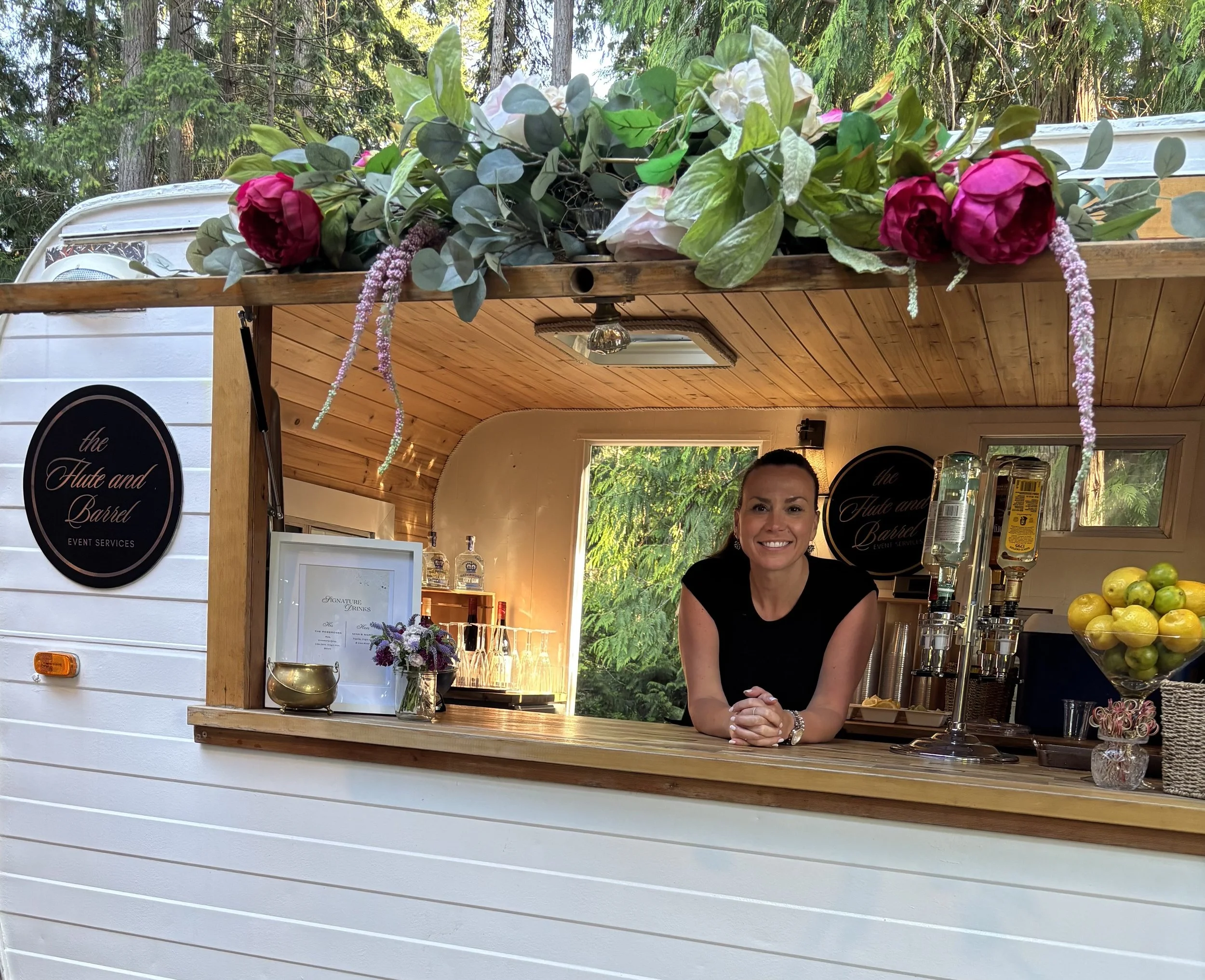 A woman with dark hair smiling behind the counter of a mobile bar service called 'The Flute and Barrel'. The bar has a wooden interior, floral decorations on top, and a window showing greenery outside.