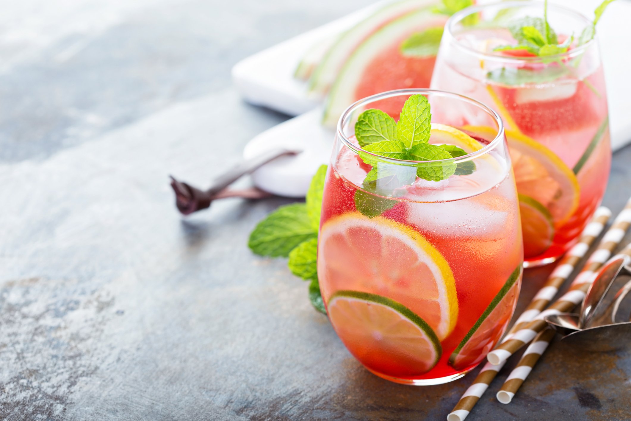 Two glasses of pink lemonade with lemon, lime slices, and mint leaves on a table with striped paper straws and a white plate of watermelon slices in the background.